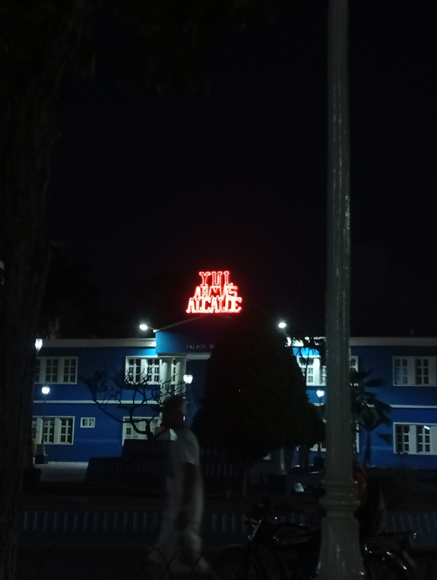 At night, a blue building with white-framed windows is seen, topped with a red neon sign that reads "YU ALCALDE". The building's facade includes pillars and a balcony. A lamppost stands in the foreground, partially obscuring the view. A person in light-colored clothing is walking on a path in front of the building. Bicycles are parked near the lamppost. Trees are visible on either side of the building, adding to the night scene in Juan Griego, Venezuela.