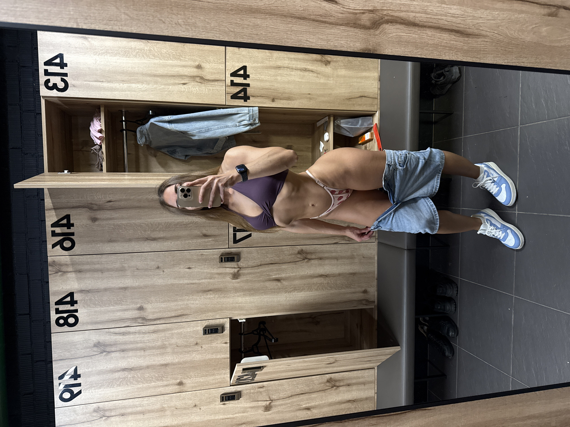In a locker room with wooden lockers, a person with athletic build is standing in front of a mirror, taking a selfie. They are wearing a purple bikini top and strawberry-patterned bikini bottoms, with denim shorts partially unzipped and lowered around their hips. The person is also wearing white sneakers with blue accents. The locker doors are open, revealing some clothing and personal items. A bench is located below the lockers, and several pairs of shoes are visible underneath it. The floor is tiled in a dark gray color, and the walls are made of dark brick. The lockers are numbered with symbols that appear to be Cyrillic letters. The overall environment suggests a gym or fitness facility.
