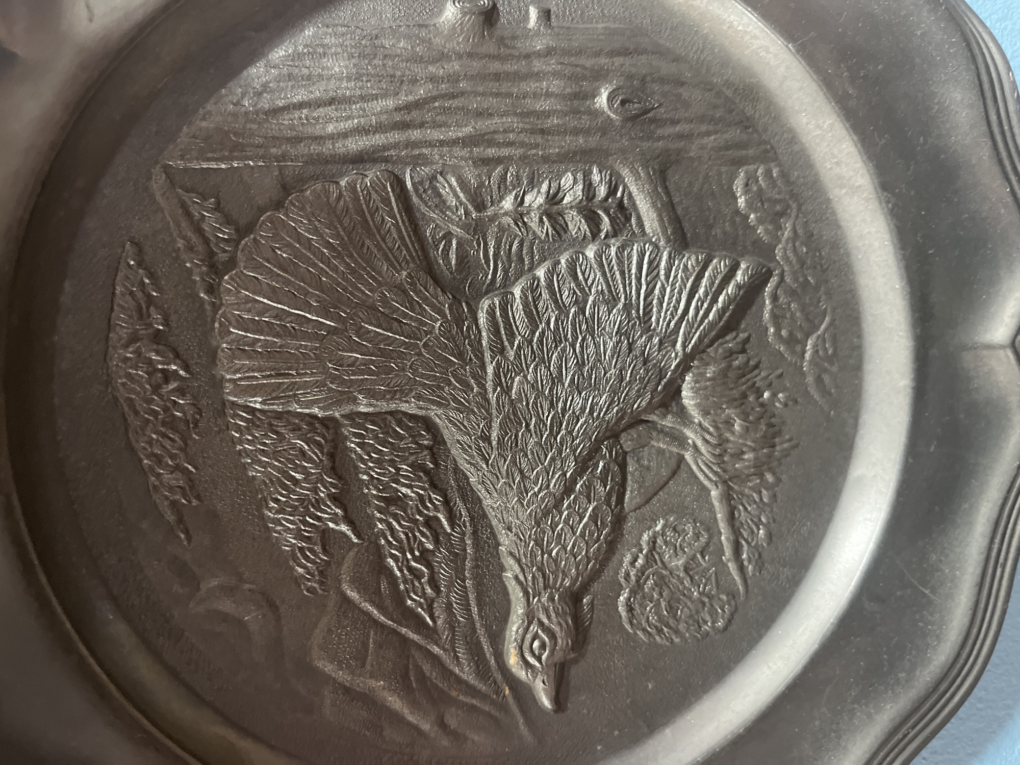 This is a detailed photograph of a circular relief plaque, likely made of pewter or a similar metal, featuring an embossed scene of a grouse in flight amidst a mountainous, forested landscape. The plaque is oval-shaped with a raised rim.
The central subject is a large grouse, depicted in mid-flight with its wings spread and fanned out, revealing intricate feather details. The bird's head is turned to the left, with its eye and beak clearly defined. It appears to be perched on a branch or a stylized tree.
The background depicts a textured mountainous terrain with stylized trees and foliage. To the right, a large tree trunk is prominently featured, with bark textures and a knot or branch visible. In the upper left corner, there's a suggestion of a crescent moon or a curved mountain ridge. The overall impression is one of a natural, wilderness setting.
The lighting emphasizes the raised details of the relief, creating shadows that add depth and dimension to the scene. The plaque exhibits some minor surface wear and subtle scratches consistent with age and handling.
This object is likely decorative and could be a souvenir, a commemorative item, or a piece of artistic craftsmanship. Without further context, its specific location in Schwäbisch Hall, Germany, cannot be determined from the image alone.