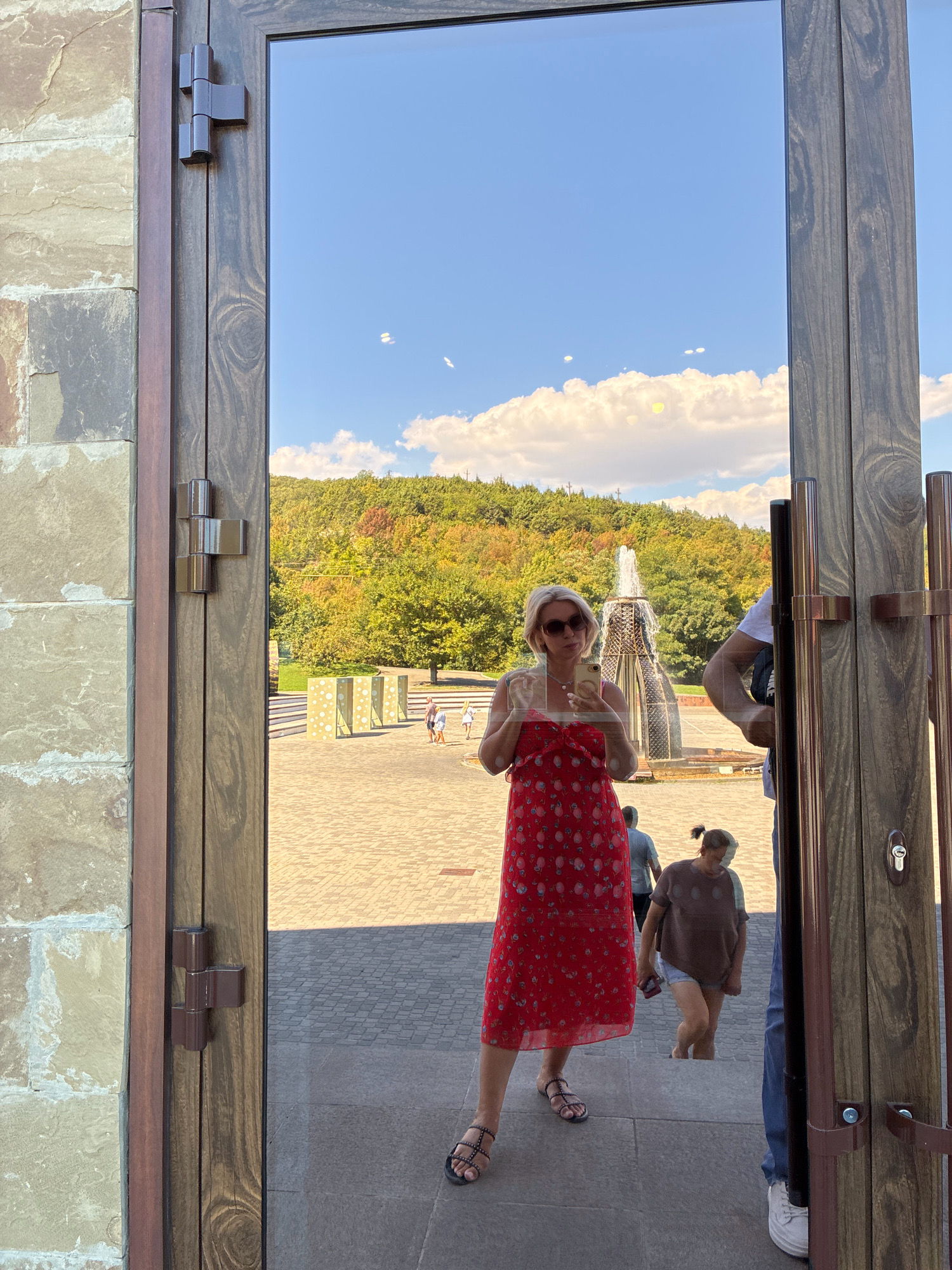 The image is a reflection in a large glass door of a woman in a red dress and sunglasses taking a selfie with her phone. She is standing on a paved area with stone walls to her left and a wooded hill with a fountain in the background. Other people are visible in the reflection, including a person in a brown shirt and shorts walking up stairs. The scene appears to be in the city of Abrau-Dyurso, Russia, likely at a public outdoor space.