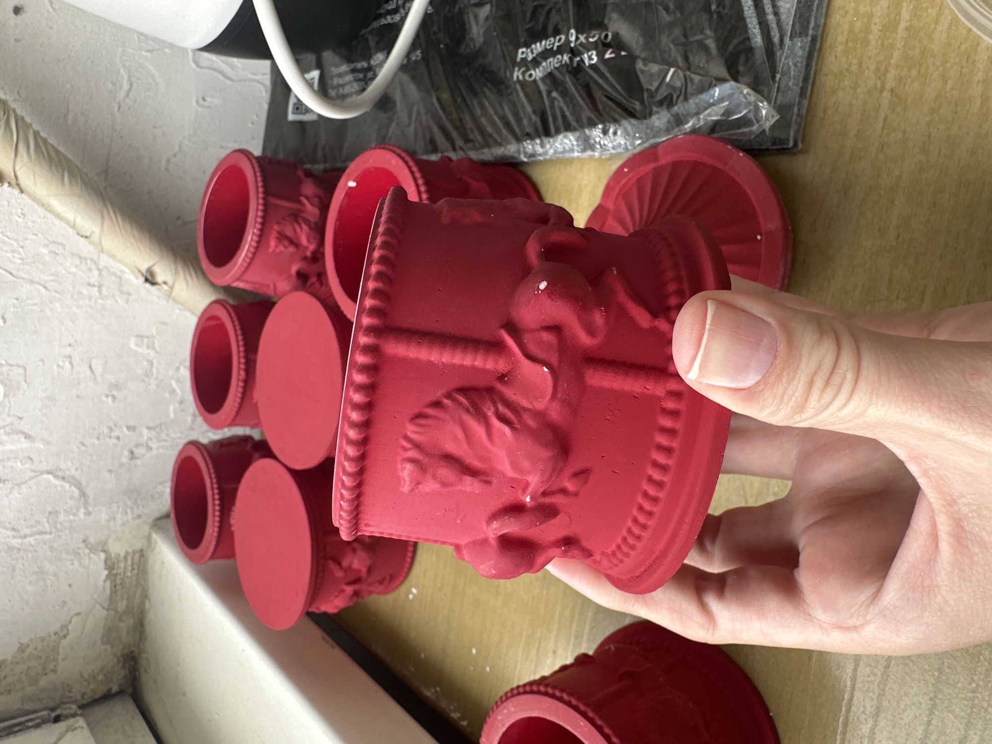 A hand holds a bright red, carousel-themed cup or holder against a blurred background. The cup features embossed horses and decorative borders. Several similar red cups and lids are visible in the background, scattered on a wooden surface. The environment appears to be indoors, possibly a workshop or storage area, with a white wall and some clutter in the background. There is no indication of the city of Vladivostok, Russia, in the image.