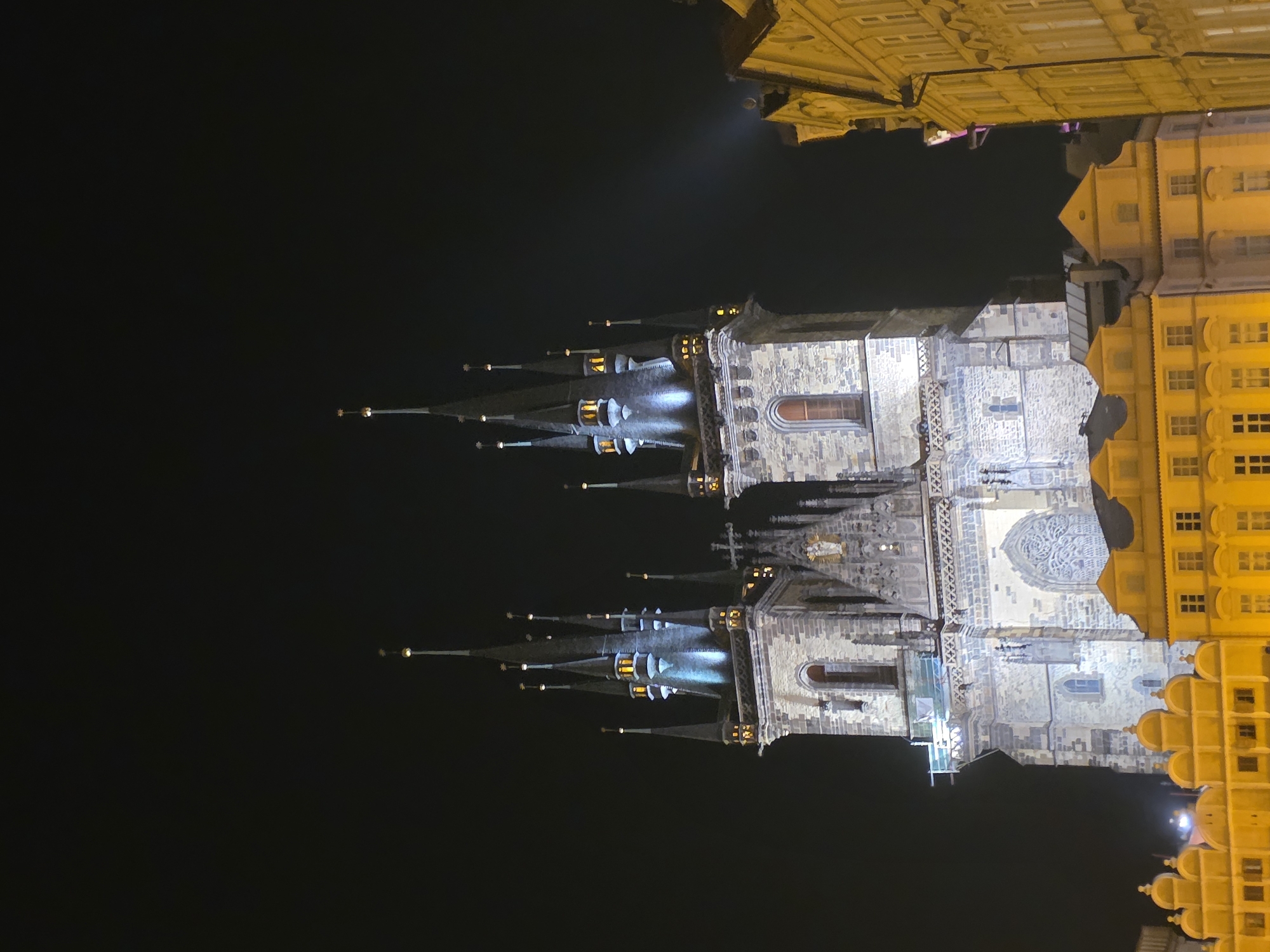 The image shows the Church of Our Lady before Týn in Prague at night. The church's spires are illuminated, and the building is adjacent to yellow buildings with multiple windows. The sky is dark.
