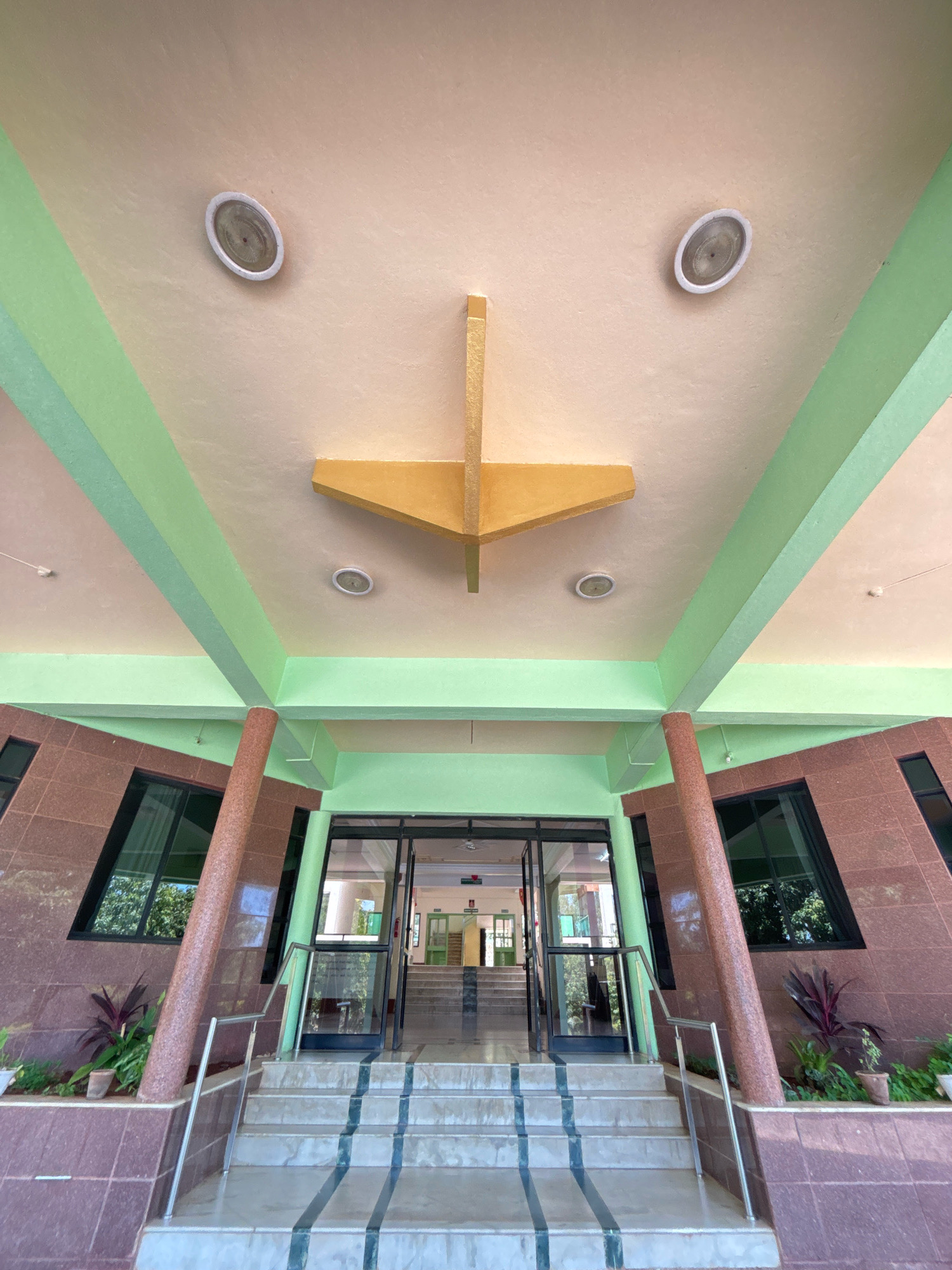 The image shows the entrance to a building, with a wide staircase leading up to glass doors. The building has a modern architectural style, with a light-colored ceiling and green-painted beams. The walls are covered in brown granite tiles, and there are large windows. Two oval-shaped light fixtures are visible on the ceiling. A decorative, yellow geometric structure is suspended from the ceiling. Plants in pots are visible on either side of the entrance. The scene appears to be a public building, possibly an office or educational institution, in Jhangatihala, India. There are no people visible in the image.
