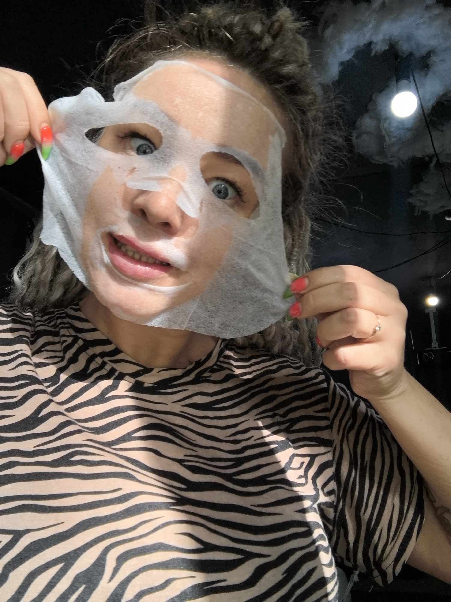 A woman is holding a white sheet mask to her face, looking playfully at the camera. She is wearing a zebra-striped shirt. Her nails are painted with a red to green gradient. The background is dark, with what appears to be decorative clouds and a light fixture. The location and the city of Noviy, Russia, cannot be determined.