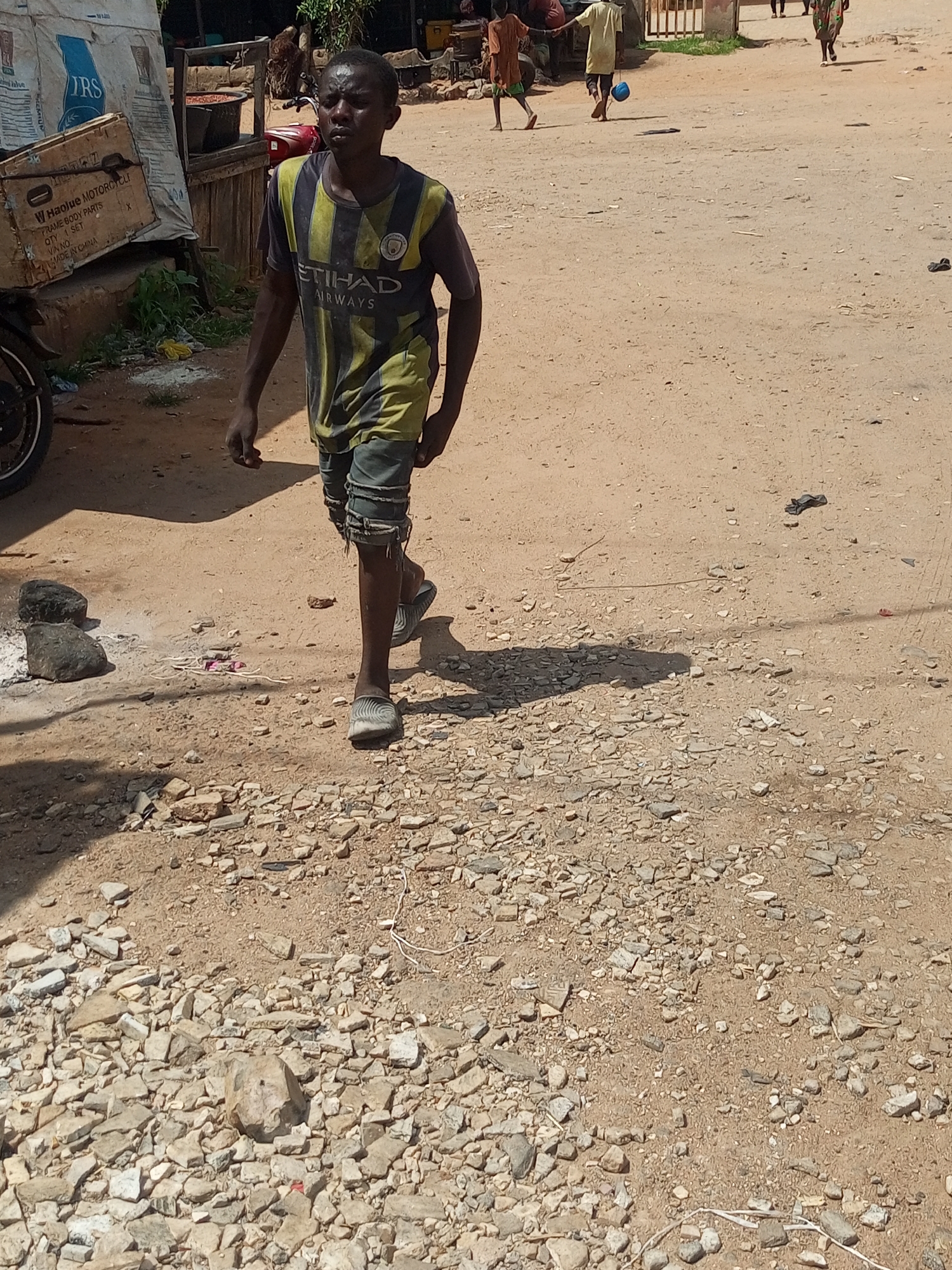 A young male wearing a yellow and black striped t-shirt with "ETIHAD AIRWAYS" printed on it, and torn denim shorts, walks on a dirt road. He is wearing grey and white striped sandals. Behind him, to the left, is a white tarp-covered object and a cardboard box with Chinese writing on it, labeled "HAOJU MOTORCYCL". To the right of the tarp, a red motorcycle is parked. Further in the background, there are several people walking, one of whom is carrying a blue bucket. The environment is sandy and rocky with scattered debris. The scene appears to be in an outdoor market or village setting.