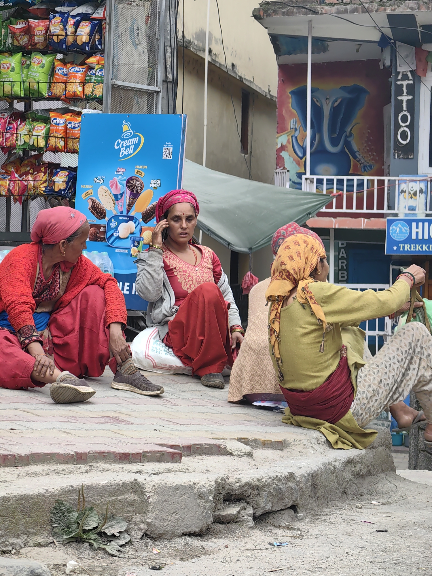 In Vashisht, India, four women are seated on a concrete ledge outdoors. One woman, on the left, wears a red shawl and red pants and looks towards another woman. The second woman, in the center, wears a pink headscarf and a pink patterned top with red pants, and is speaking on a mobile phone while looking down. She is sitting on a white sack. To the right, a third woman, wearing a yellow headscarf and a light green tunic with a red sash, is looking towards the right. Her legs are covered by patterned pants. Behind her, a fourth woman is partially visible, wearing a cream-colored patterned garment. In the background, shelves are stocked with packaged snacks and a blue sign displays ice cream advertisements. A colorful mural of a blue elephant is painted on the wall of a building to the right. The environment appears to be a street scene with buildings and signage indicating commercial activity.