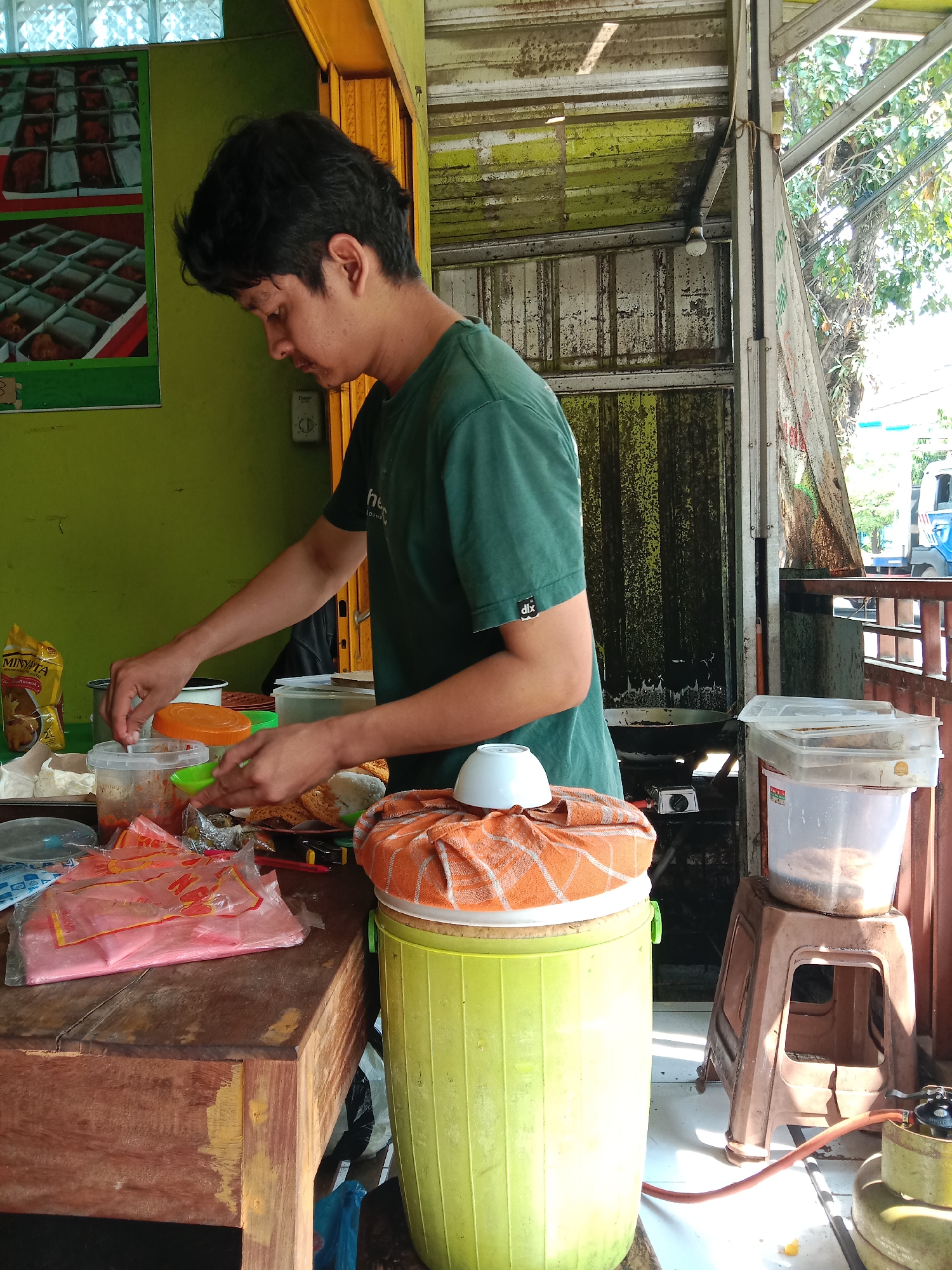 This image captures a man preparing food at a rustic street food stall in Sekopek Wetan, Indonesia, under bright daytime lighting. The scene suggests a bustling, local eatery.
The central subject is a man, likely in his 20s or 30s, seen in profile from the chest up, facing left. He has dark, medium-length hair and is wearing a dark green t-shirt with a small white "dlx" logo on the left sleeve. His hands are actively engaged in food preparation, placed over a clear plastic container holding a red or orange sauce, while he holds a small green dish.
The foreground features a worn wooden counter, upon which various items are arranged. These include several clear plastic containers with lids (some open), a yellow pouch labeled "MINYAK SUNNAH" (cooking oil), and multiple plastic bags, including a pink one with red text. Beside the counter, a light green plastic barrel, possibly a cooler or storage container, is draped with an orange-patterned cloth and topped with a small white bowl. To the right, a brown plastic stool holds another large clear plastic container. On the ground, a yellow gas cylinder with a red hose connects to a small gas stove, indicating active cooking.
The background shows the structure of the stall. The left wall is painted bright green and features two framed posters depicting various food items in square boxes. The upper sections of the stall and roof are constructed from corrugated metal, showing signs of rust and wear. An exposed light bulb hangs from the roof. Bright sunlight streams in from the right, illuminating parts of the scene and casting shadows. Beyond the stall's opening on the right, green foliage, trees, and a partial view of a vehicle are visible, confirming the outdoor setting. The overall impression is of a functional and well-used food vendor preparing meals.