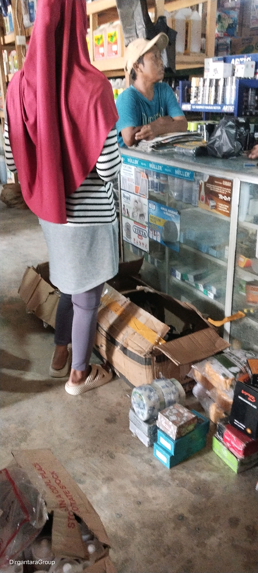 A medium shot shows two individuals, one in the foreground wearing a red hijab and a black and white striped shirt with a grey skirt, and another in the background wearing a light-colored baseball cap and a blue t-shirt. They are standing in what appears to be a retail store, possibly a pharmacy or a general store, with shelves filled with various products. The floor is concrete and littered with cardboard boxes, one of which is open and spilling its contents. In the background, a glass display case is visible, stocked with items. The overall lighting suggests an indoor environment, likely during daylight hours. The scene is suggestive of a transaction or interaction between the two individuals, with the person in the background appearing to be an employee or vendor.