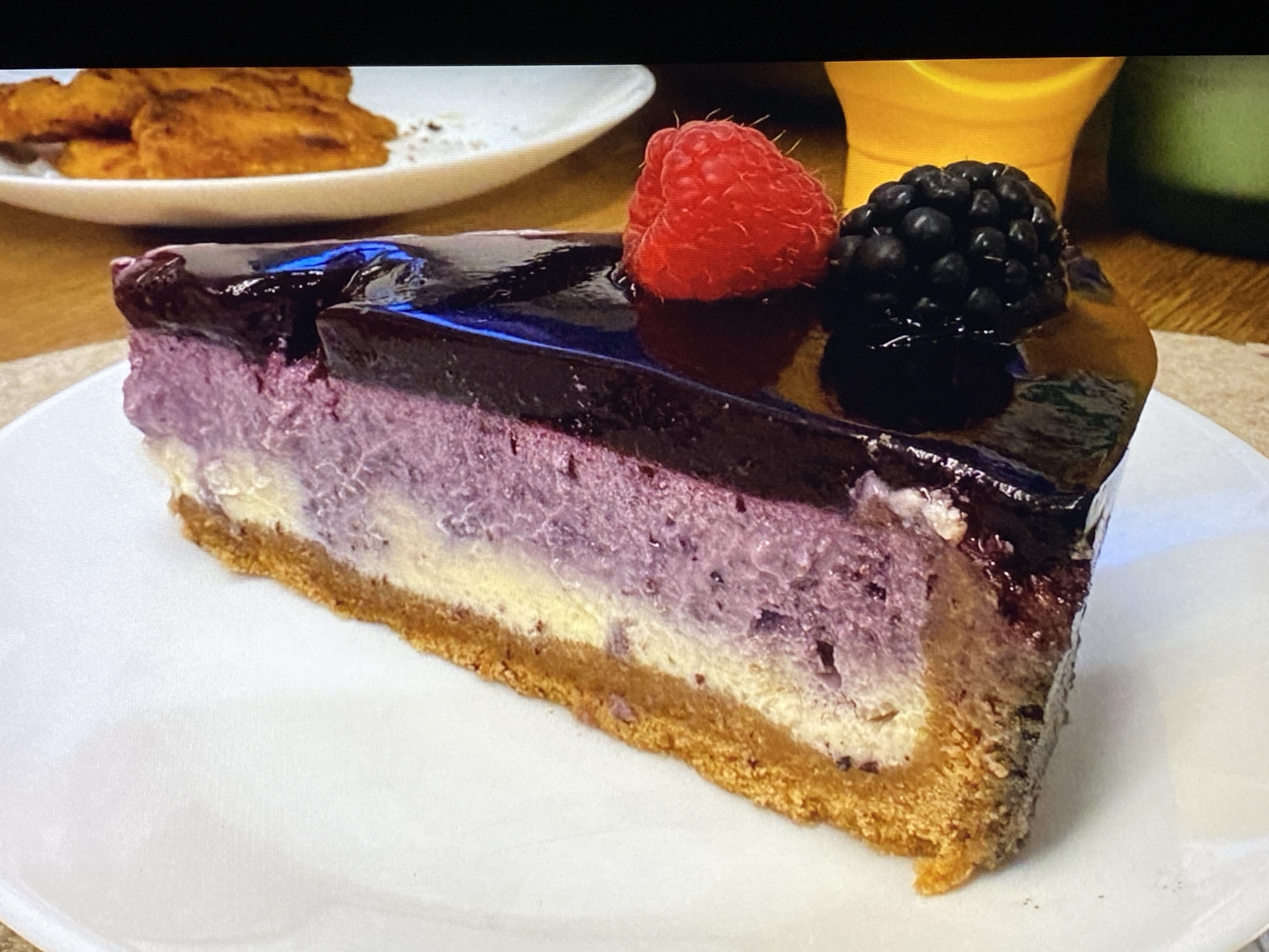 A slice of blueberry cheesecake sits on a white plate. The cheesecake has a thick graham cracker crust, a layer of creamy white filling, and a purple-hued blueberry filling. It is topped with a dark purple glaze and garnished with a fresh raspberry and a blackberry. In the background, out of focus, is a white plate with some orange food items, a yellow object, and a green object. The scene appears to be indoors, possibly in a restaurant or home setting. There is no information in the image to suggest a specific location in Purkersdorf, Austria.