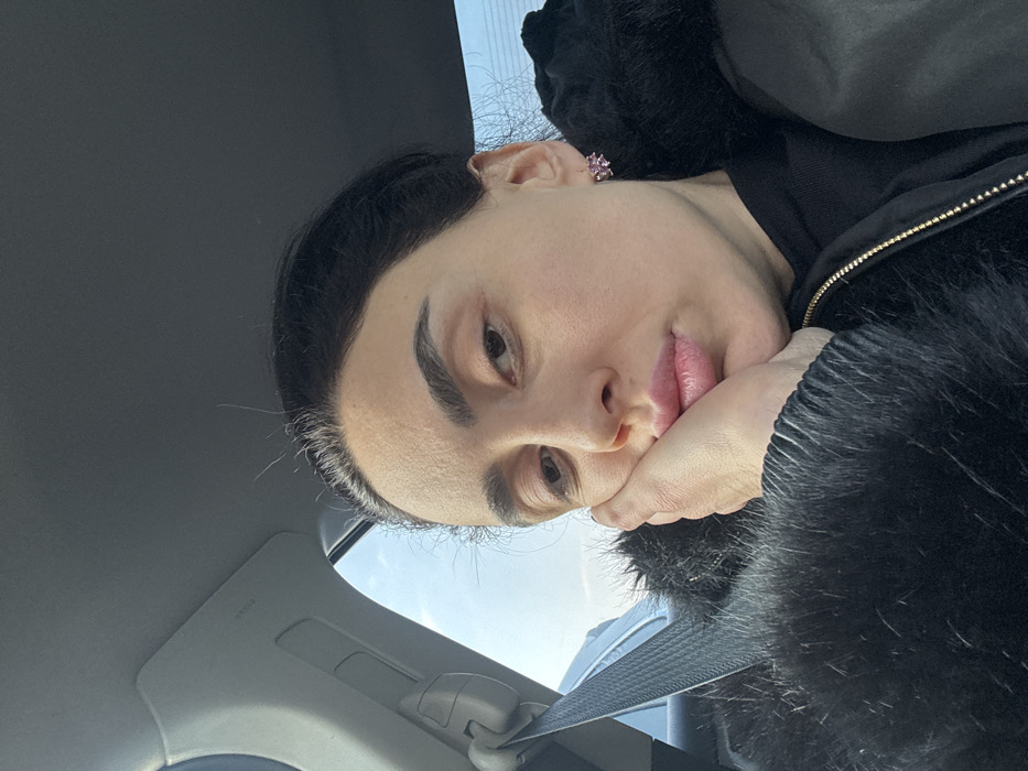 A selfie shows a person resting their face on their hand, wearing a black furry jacket and a pink earring. The person appears to be inside a car, with the car's ceiling and a seatbelt visible in the frame. Outside the car's window, a glimpse of the sky can be seen. The overall perspective is angled, looking upward towards the person.