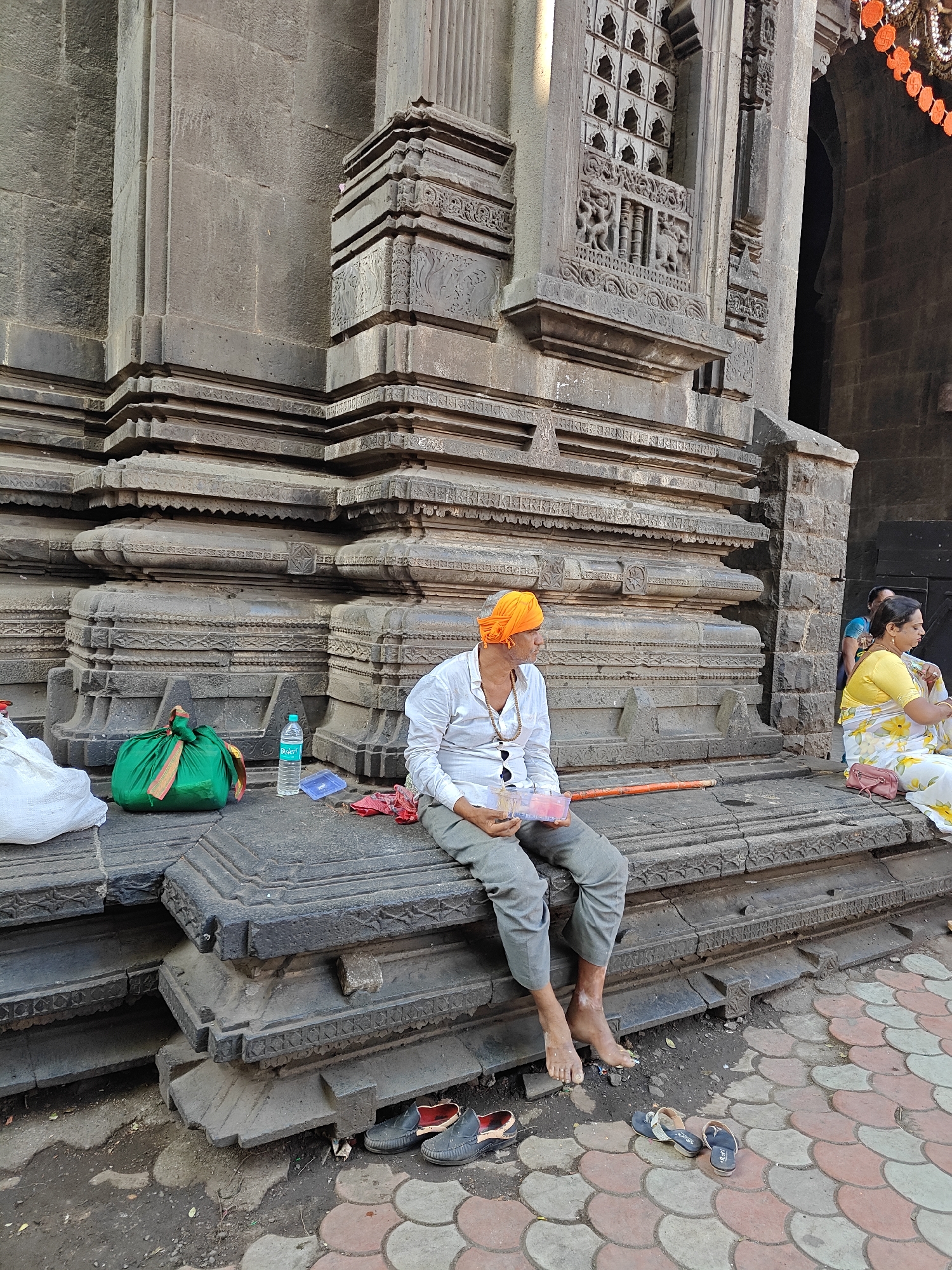 A medium shot shows an Indian man with bare feet sitting on steps in front of a stone building. The man is wearing an orange turban, gray pants, and a white long-sleeved shirt. He is holding a clear plastic container and looking to his right. Next to him are a green bag, a water bottle, and a red cloth. Pairs of shoes and sandals are on the ground below the steps. To the man's right, a woman in a yellow and white floral dress is seated. The building behind them is made of gray stone with carved details. The location is Kolhapur, India.
