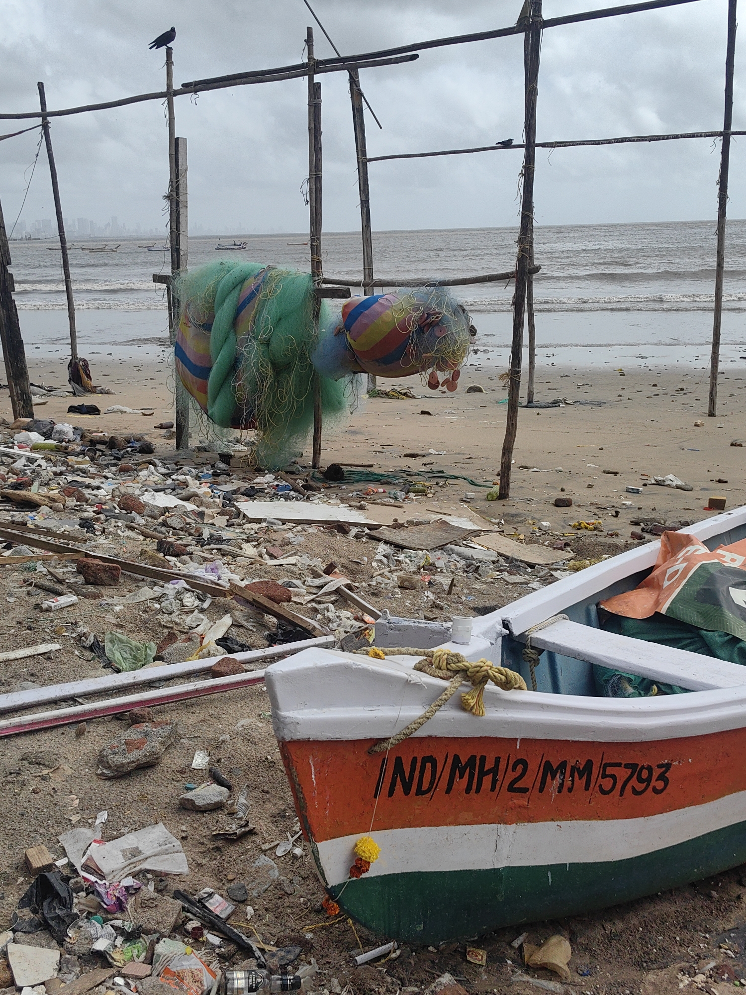 The image depicts a cluttered beach scene in Mumbai, India, under an overcast sky. In the foreground, a small fishing boat with the registration "IND/MH/2/MM5793" is pulled onto the shore. The boat is painted with orange and green stripes on its hull and has a yellow garland with marigold flowers attached to its side. The beach itself is strewn with a significant amount of debris, including plastic waste, wood fragments, fishing nets, and discarded items. Behind the boat, a large, colorful fishing net is draped over a makeshift wooden structure. In the background, the Arabian Sea is visible, with gentle waves rolling onto the shore. Several small boats are scattered on the water, and a hazy city skyline, likely representing Mumbai, is visible in the distance. A dark bird is perched on a wooden pole in the midground. The overall impression is one of neglect and environmental pollution on the beach.