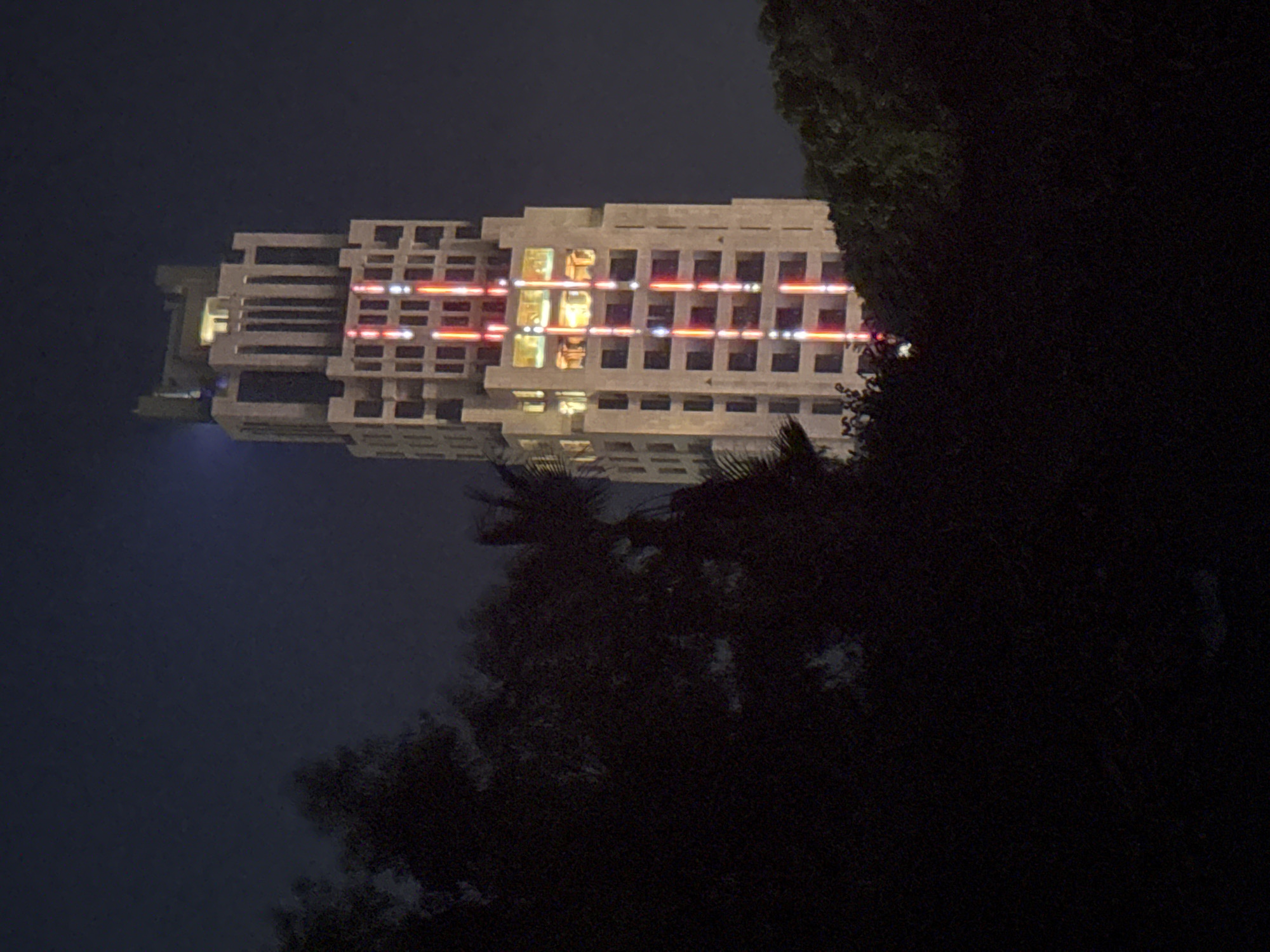 The image shows a tall, modern building in the city of Parekklisia Community, Cyprus, at night. The building is illuminated with vertical red and white lights, creating a striking visual effect against the dark sky. Trees and foliage partially obscure the lower part of the building, framing the scene with dark shadows. The top of the building features a distinctive architectural design with vertical columns and a lighter-colored structure at its peak. A faint blue light is visible in the upper left portion of the sky. The overall impression is one of an urban landscape at night, characterized by artificial lighting and the presence of surrounding nature.
