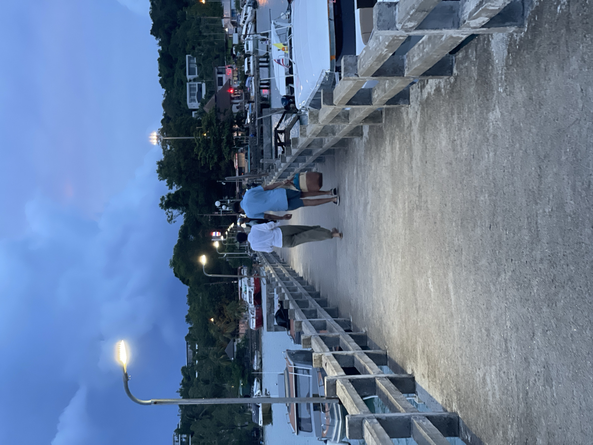 The image shows a paved pier at dusk in Pa Khlok, Thailand. Two people are walking away from the camera along the pier, with one person wearing a light blue short-sleeved shirt and dark shorts, and the other wearing a white short-sleeved shirt and khaki pants. The person in blue is carrying a brown bag. The pier is lined with concrete railings and streetlights that are illuminated. To the left of the pier, boats are docked in the water, and beyond the boats, lush green trees and buildings are visible on a hillside. The sky is a deep blue with some clouds, indicating the transition from day to night. The overall mood of the image is calm and serene, suggesting a peaceful evening stroll. There is no visible text in the image.