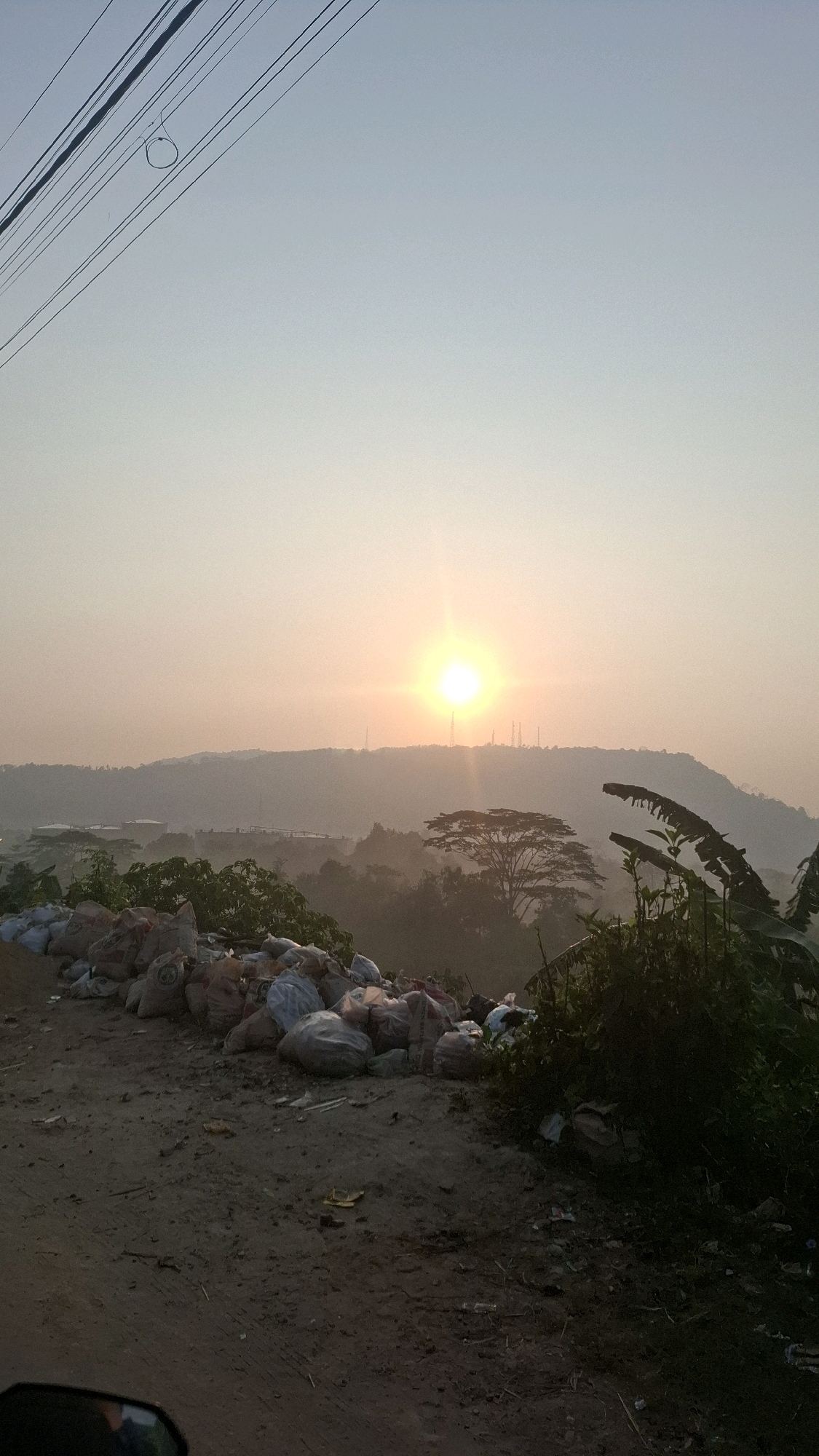This is a vertical, medium-shot photograph of a sunset over a hazy landscape in Bandar Lampung, Indonesia. The foreground is a dirt path with a large pile of garbage bags on the left, and dense foliage on the right. In the middle ground, there are buildings and trees. The background consists of rolling hills and a bright, hazy sun. Overhead, there are power lines, one with a circular loop. The image appears to be taken from a moving vehicle, as indicated by a dark reflection in the lower left corner.