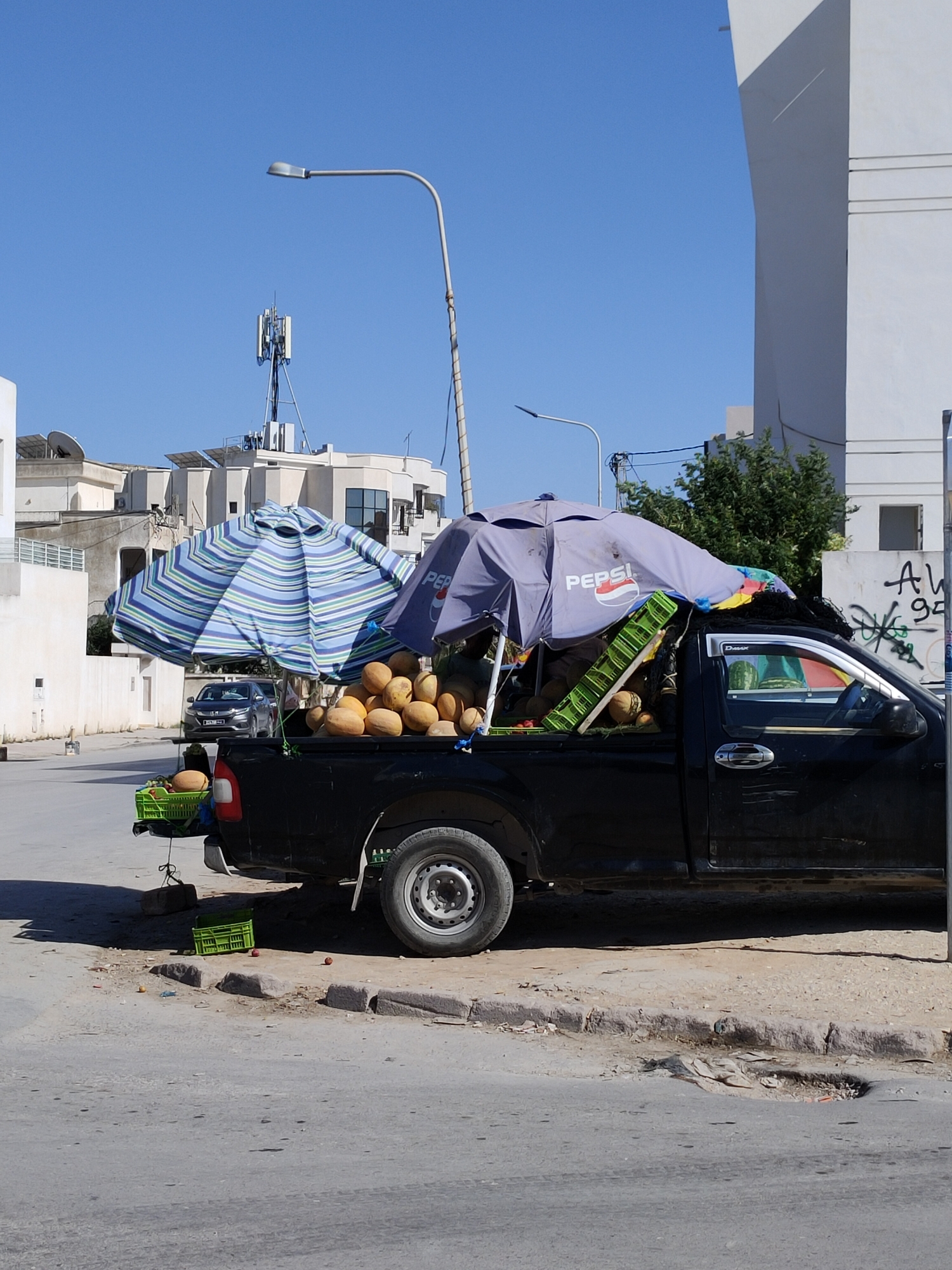 The image depicts a black pickup truck parked on a street in Ariana, Tunisia, laden with a variety of fruits and produce. Two large umbrellas, one striped blue and green and the other purple with a Pepsi logo, are erected over the truck bed, providing shade for the goods. The truck is filled with what appear to be melons, some of which are contained within green plastic crates. Additional green crates are visible on the side and back of the truck, and some loose fruits are scattered on the ground near the curb.
In the background, residential buildings with flat roofs and satellite dishes are visible under a clear blue sky. A street light and a telecommunications tower are also present. The street appears to be paved, with a concrete curb and a pothole in the road. The overall scene suggests a street vendor operating from a vehicle. There are no people visible in the frame.