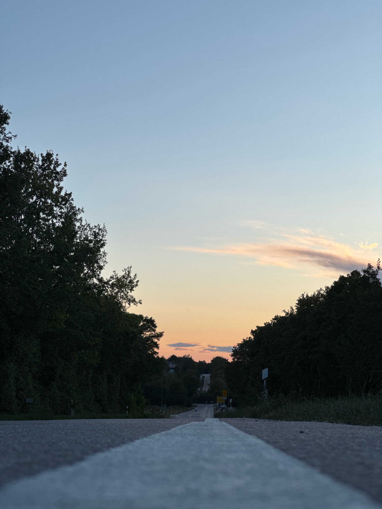 A low-angle, wide shot captures a paved road with a prominent white line in the foreground. The road recedes into the distance, flanked by dense, dark green trees on either side. The sky above transitions from a pale blue at the top to soft hues of pink and orange near the horizon, indicative of a sunset. In the background, there are faint structures and more trees, suggesting a semi-rural or suburban setting. The image appears to be taken in Wettstetten, Germany, during the evening. There are no people or visible actions in the frame.