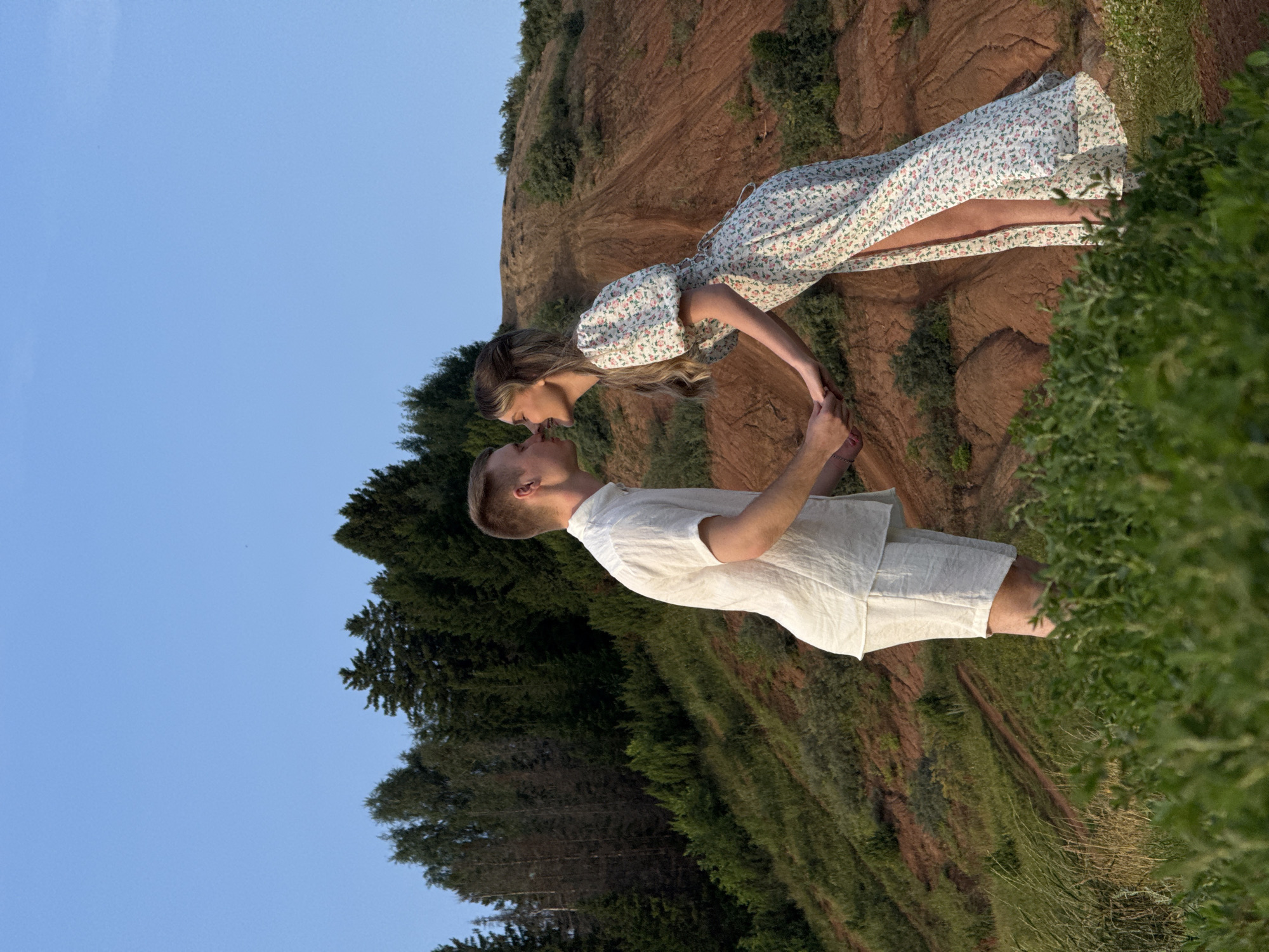 A couple is captured in a romantic moment, about to kiss on a grassy slope in Olkhovka, Russia. The man, wearing a white linen shirt and shorts, stands facing the woman. The woman, in a floral print maxi dress with puff sleeves, leans towards him, holding his hand. They are positioned in front of a backdrop of a steep, reddish-brown hillside dotted with sparse greenery, and a dense forest of dark green trees behind it, under a clear blue sky. The foreground is filled with lush green vegetation, partially obscuring the lower half of the couple.
