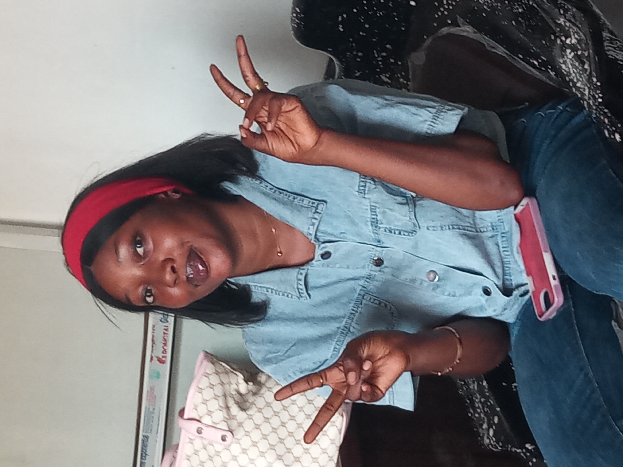 A young Black woman is depicted in a casual indoor setting in Sabon Tasha, Nigeria. She is seated or leaning, looking towards the camera with a playful expression, her mouth slightly open. Both of her hands are raised, forming peace signs. She has long, straight black hair styled with a red headband.
She is wearing a light blue, short-sleeved denim shirt with snap buttons, and dark blue jeans. Accessories include a thin gold-colored necklace, a gold-colored bracelet on her left wrist, and several rings on her fingers (on the right index, right ring, and left ring fingers). A pink phone case is visible, tucked into her right front jeans pocket.
The background features a plain white wall. To her left, a light-colored handbag with a patterned design is visible. Behind her, a dark patterned surface, possibly part of a chair or sofa, can be seen. On the white wall behind her head, a rectangular object or poster is present, with partially visible text that reads "DIGITAL Girl". The lighting suggests it is daytime. The overall mood of the image is casual and cheerful.