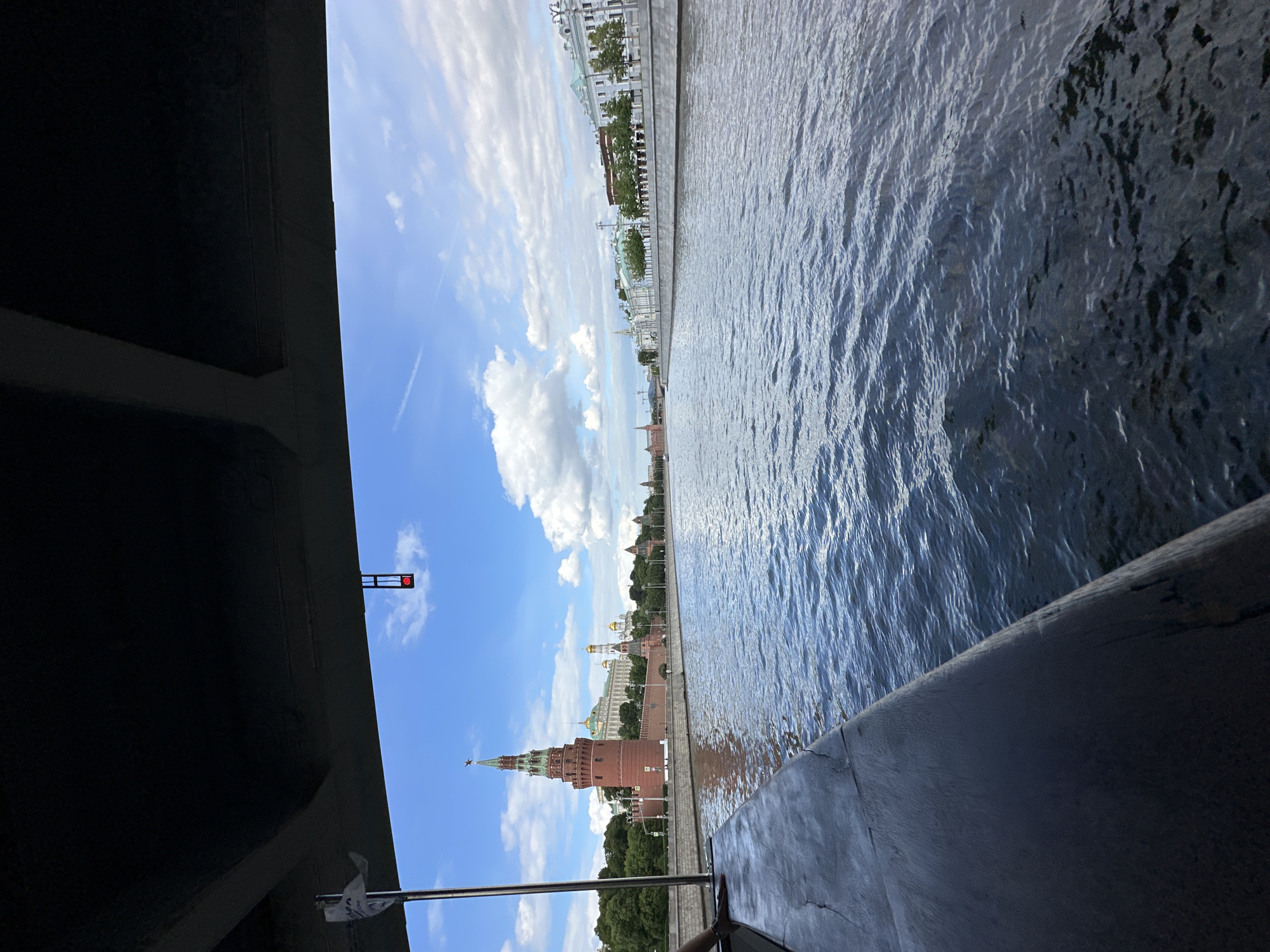 The image shows a view from under a bridge or structure in Moscow, Russia, looking out towards the Moscow Kremlin. The Moscow River is prominent in the scene, with its surface rippled by the wind. The Kremlin towers, including the Spasskaya Tower with its red star, are visible against a backdrop of a blue sky with scattered white clouds. A building or promenade lines the river on the opposite side. The view is partially obscured by a dark structural element, either part of the bridge or another structure. A small red light is seen on the edge of this structure. A piece of fabric or paper is attached to a railing or fixture.