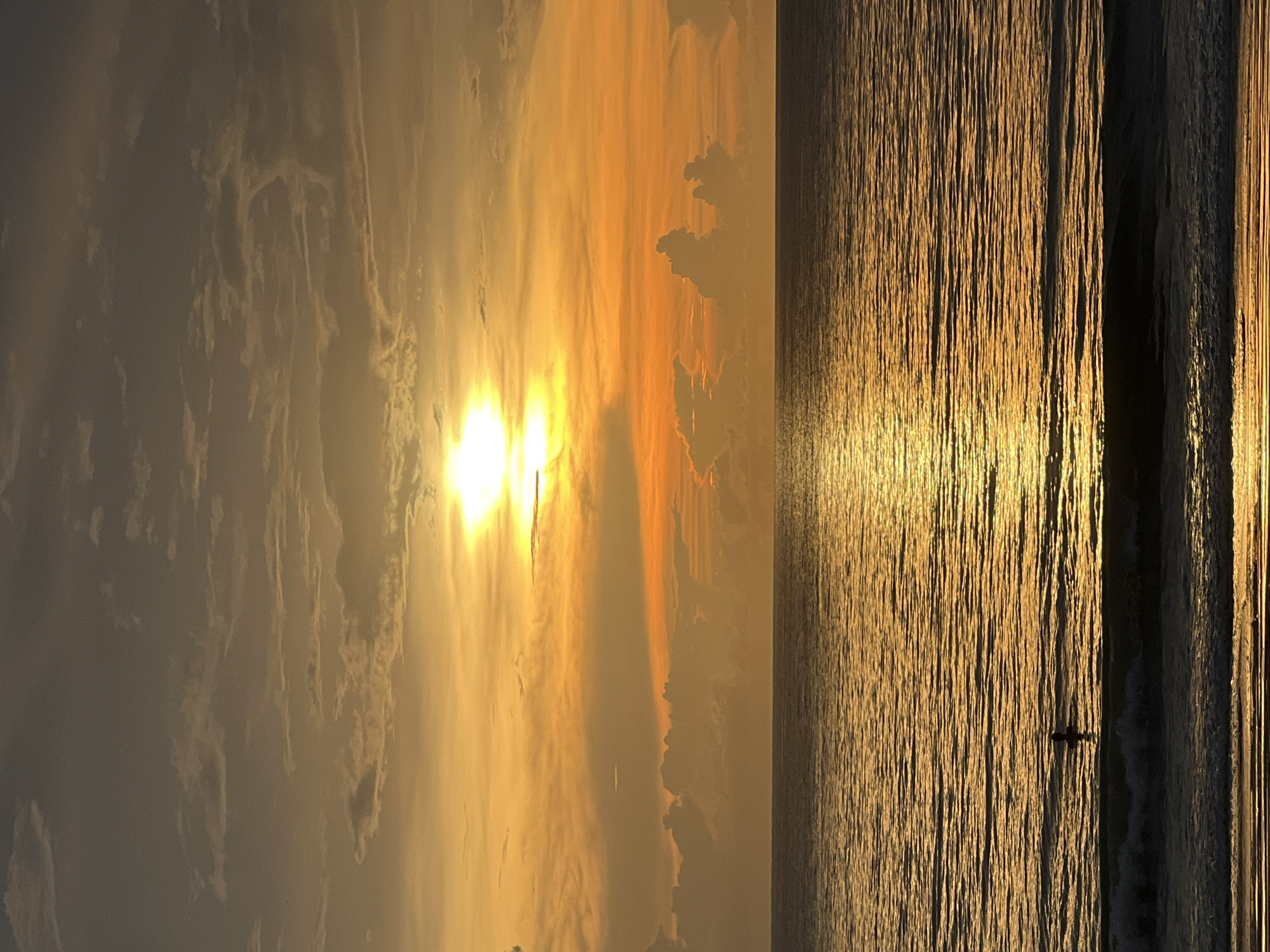 The image shows a golden sunrise or sunset over the ocean. The sky is filled with clouds, and the sun is shining brightly, partially obscured by clouds. The ocean reflects the sunlight, creating a shimmering effect. In the ocean, there is a silhouette of a person on a paddleboard. The location is unknown.