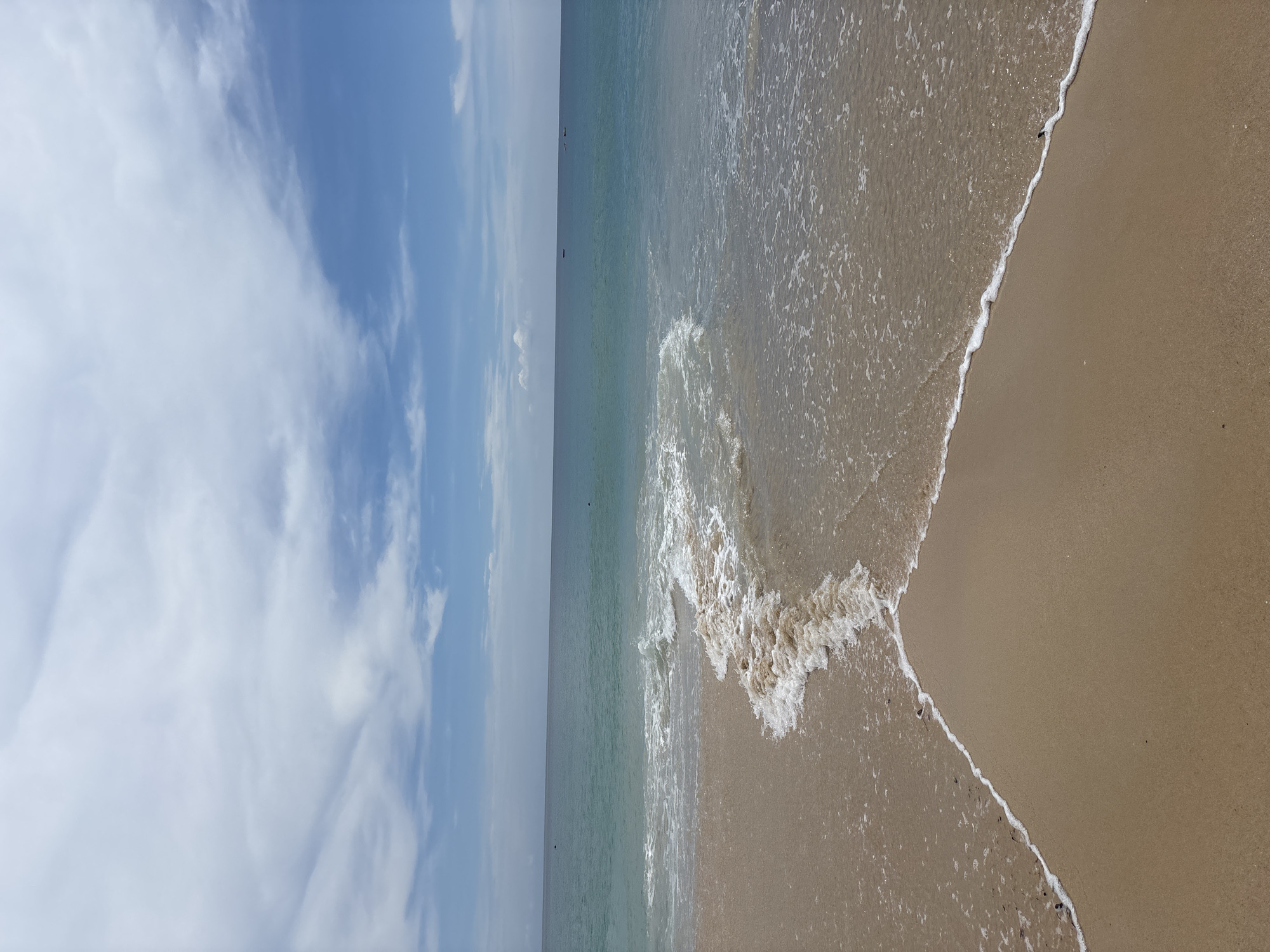 The image shows a beach in Choeng Thale, Thailand. The scene depicts a sandy beach with waves gently lapping the shore. The sand is a light tan color. The ocean water transitions from a darker hue further out to a lighter, foamy texture closer to the shore. The sky is blue with patches of white clouds. Two small boats are visible in the sea.