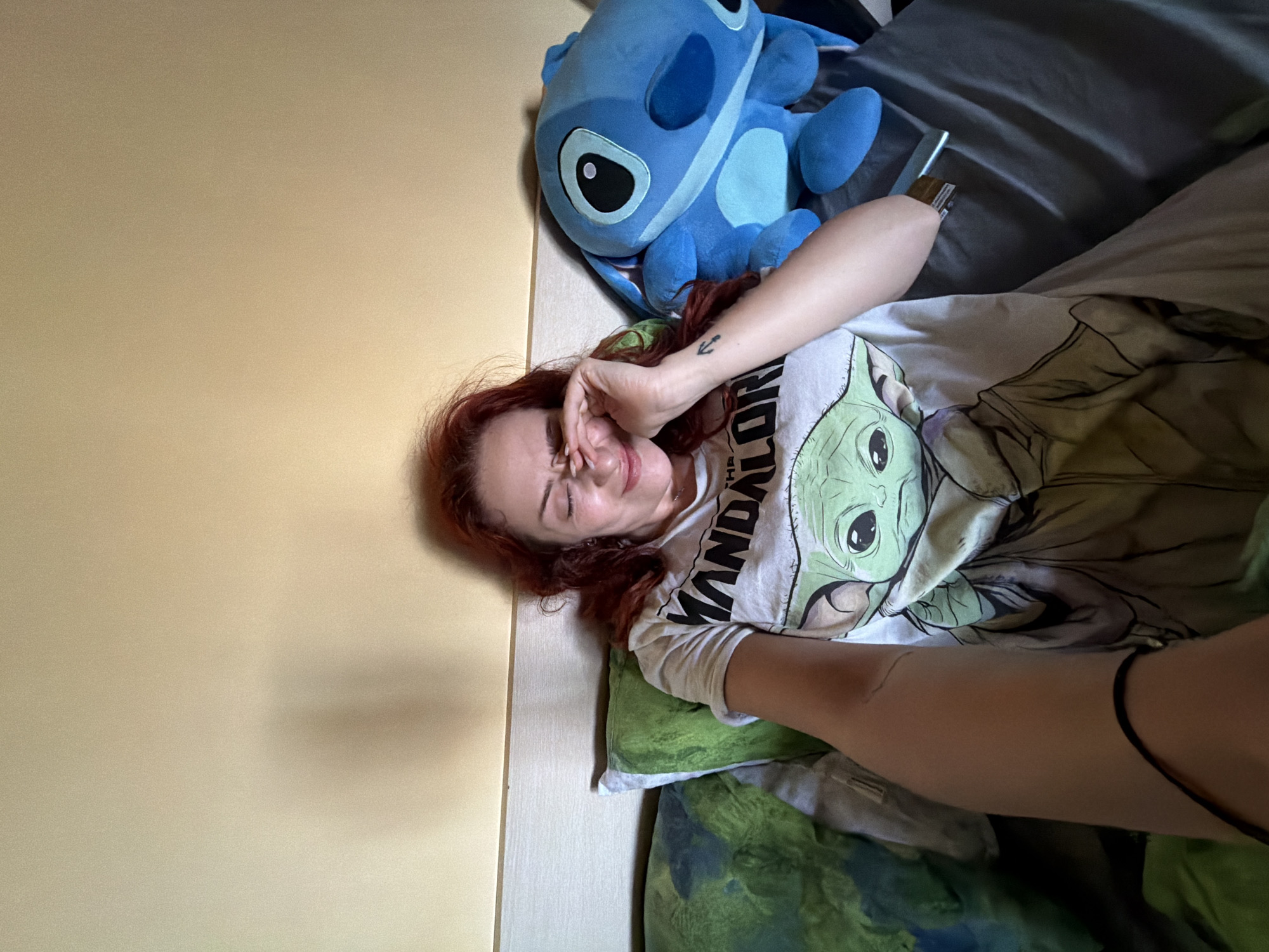 A person lies on a bed in a room. The person has reddish hair and is wearing a white t-shirt with an image of "Grogu" (Baby Yoda) and the words "The Mandalorian" on it. They are partially covered by bedding. A blue Stitch plush toy is next to the person's head. A cell phone rests on the bed. The location is not discernible from the image.