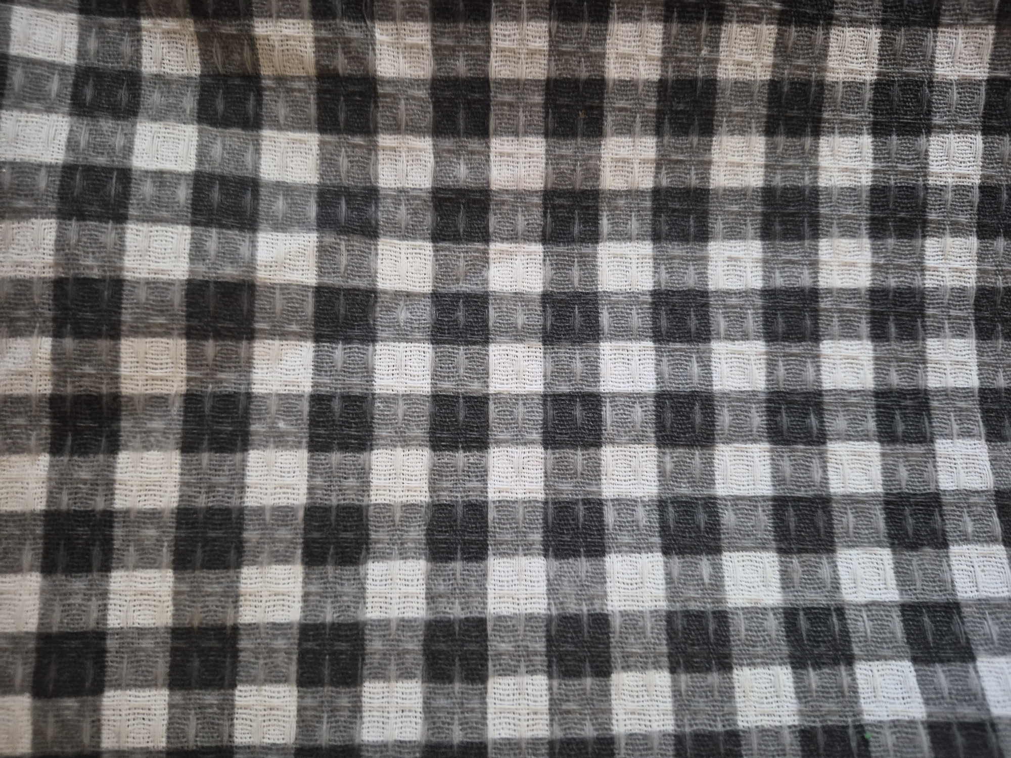 This close-up, top-down image presents a textile featuring a distinct checkered or gingham pattern. The fabric primarily consists of three shades: off-white, dark gray (almost black), and a transitional mid-tone gray. The pattern is formed by an arrangement of squares, with solid off-white blocks alternating with solid dark gray blocks. The mid-tone gray areas are created by a dense weave of both off-white and dark gray threads, resulting in a lighter-toned checkerboard within the larger pattern.
A prominent detail is the fabric's texture, which exhibits characteristics of a waffle weave or similar patterned weave. This technique creates small, raised square-like formations across the surface, giving the material a three-dimensional depth and a tactile quality. These raised elements cast subtle shadows, enhancing the visual texture.
The lighting is even and soft, suggesting either diffused natural light or gentle artificial illumination, as there are no harsh shadows external to the fabric itself. The entire frame is filled with the fabric, emphasizing its pattern and intricate weave. The material appears clean and well-maintained.
There are no people, other objects, or specific activities depicted in the image. No visible text is present. The image provides no discernable cues regarding the time of day, weather, or specific location, apart from the provided context of Yerevan, Armenia, which is not visually supported by the content.