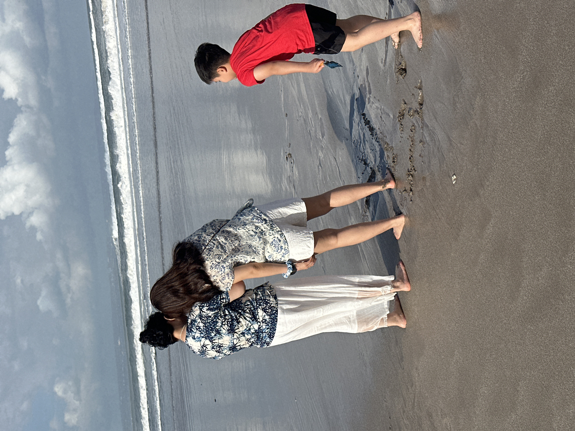 Three individuals are observed on a sandy beach in North Kuta, Indonesia. In the foreground, two individuals, likely adults, are positioned facing the ocean. One is wearing a white garment with a blue and white patterned top and a long white skirt. The other is wearing a similar patterned top and white shorts. They appear to be conversing or observing something together. To the right, a young boy in a red t-shirt and black shorts is facing away from the adults, looking down at the sand and holding a small blue object in his right hand. The beach is wet, indicating recent tide action. The ocean in the background features gentle waves and a cloudy sky.