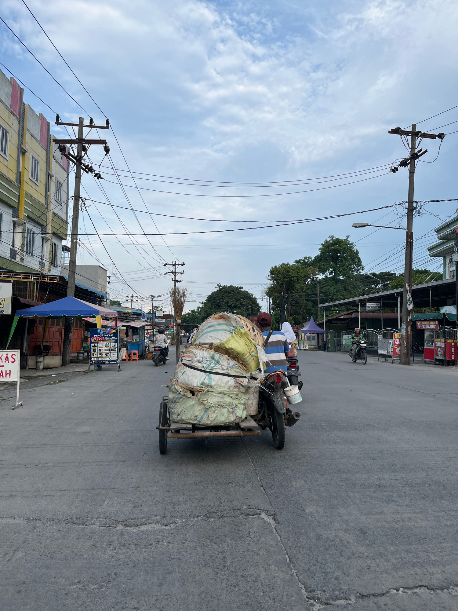 This is a street-level, outdoor, eye-level, vertical, medium shot taken in the City of Medan, Indonesia. The scene depicts a street with buildings on either side, utility poles with extensive wiring, and a cloudy sky. In the foreground, a three-wheeled motorcycle, a "becak motor," is carrying a large, bulky load wrapped in light-colored sacks. The motorcycle has two people on it; the driver in front and a passenger behind. They are moving away from the camera. In the mid-ground, other motorcycles and pedestrians are visible, as well as several small stalls or shops with awnings. The road surface shows some cracks and wear. The buildings on the left appear to be multi-story, possibly residential or commercial. The overall impression is of a busy, everyday street scene in an urban area.