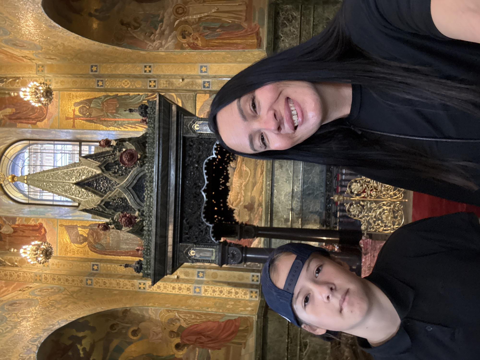 A medium shot shows a woman and a young person in the foreground, with the background of an ornately decorated church interior. The woman, on the right, has long, dark hair and is smiling broadly, with her head tilted slightly. She is wearing a black t-shirt. The young person, on the left, wears a dark blue baseball cap and a black polo shirt. They are looking forward with a neutral expression.
The background features gold-adorned walls with religious murals and intricate architectural details. A large, dark, ornate structure, possibly an altar or iconostasis, is visible behind the two individuals. The overall impression is a tourist snapshot taken inside a place of worship, characterized by its rich artistic and religious decoration.