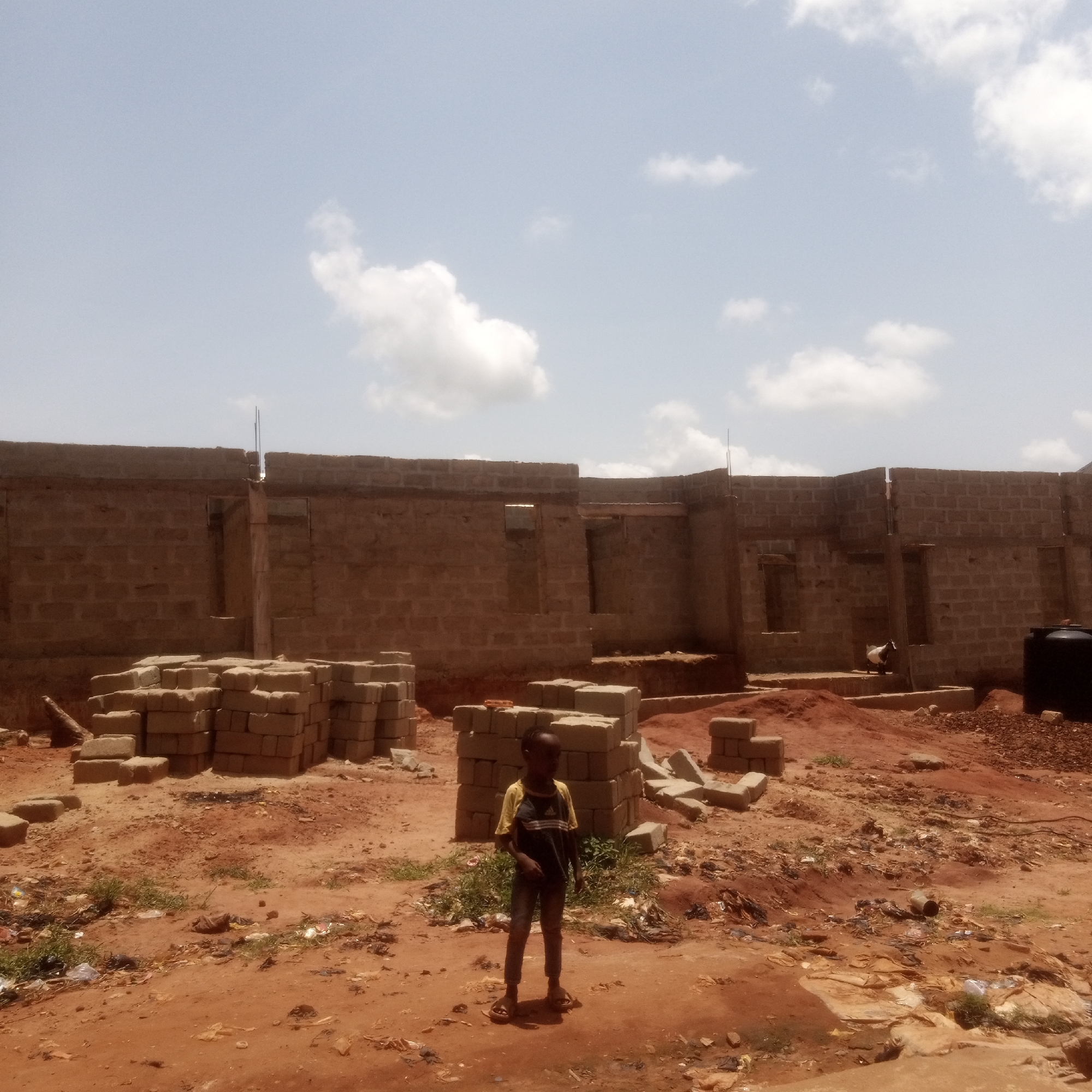 The image depicts a construction site in Ukwu-Abwa, Nigeria. An unfinished brick building stands in the background. Piles of bricks are scattered in the foreground. A young boy wearing a yellow-and-black shirt and sandals stands in the center of the frame. The ground is predominantly red soil with patches of vegetation and scattered debris. The sky is visible and features a few white clouds. A water tank and a white goat are partially visible near the building.
