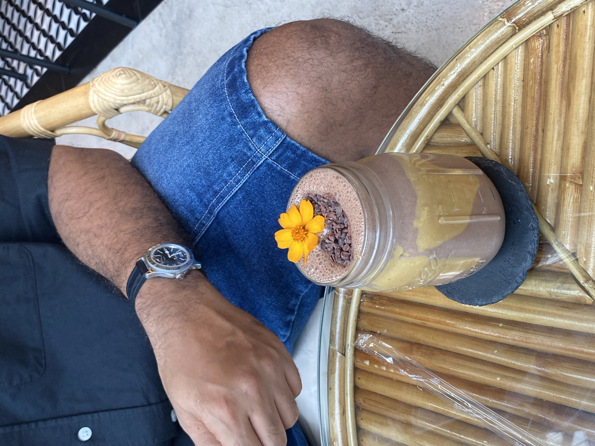 A high-angle shot shows a person seated, presumably at a cafe, with a smoothie decorated with a yellow flower. The person wears a black short-sleeved shirt, blue denim shorts, and a watch with a black band on their left wrist. The smoothie is served in a glass jar topped with chocolate nibs and has a two-toned color. It rests on a small black coaster placed on a rattan chair. The location is suspected to be in Bangkok, Thailand, but the specific details supporting this determination are not immediately apparent in the image. The focus is primarily on the person's arm and the smoothie.