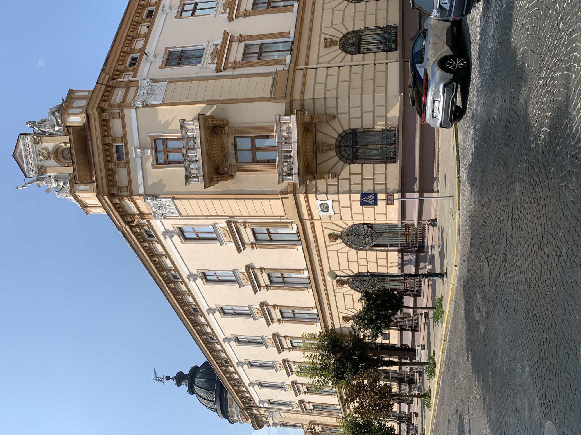 The image shows a view of a building and a street in Kolomyia, Ukraine. The building is a large, ornate structure with a light yellow facade, brown trim, and decorative architectural elements. It features multiple stories, arched windows, balconies, and statues on its roof. A dark dome with a weather vane is visible on one side of the building. The street in front of the building is paved with cobblestones. A silver car is parked on the side of the street, facing away from the camera. There are trees planted along the sidewalk in front of the building. The sky is clear and blue.