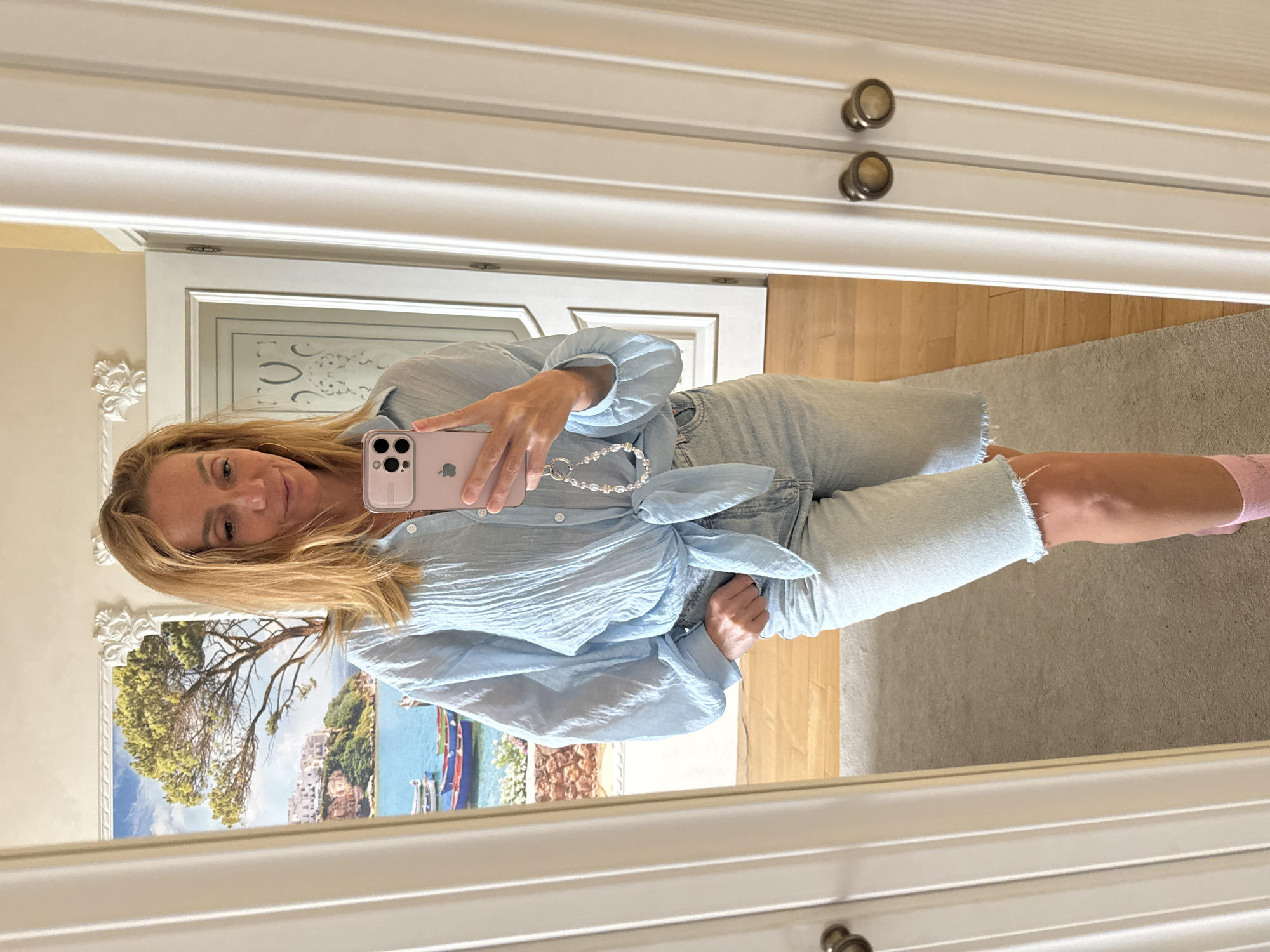 A woman with blonde hair stands in front of a mirror, taking a selfie. She is wearing a light blue, crinkled blouse tied at the waist and light wash denim shorts. A beaded phone charm hangs from her phone. In the background, a mirrored closet door reflects a painting of a coastal village with boats and a tree. The floor is a mix of wood and carpet.