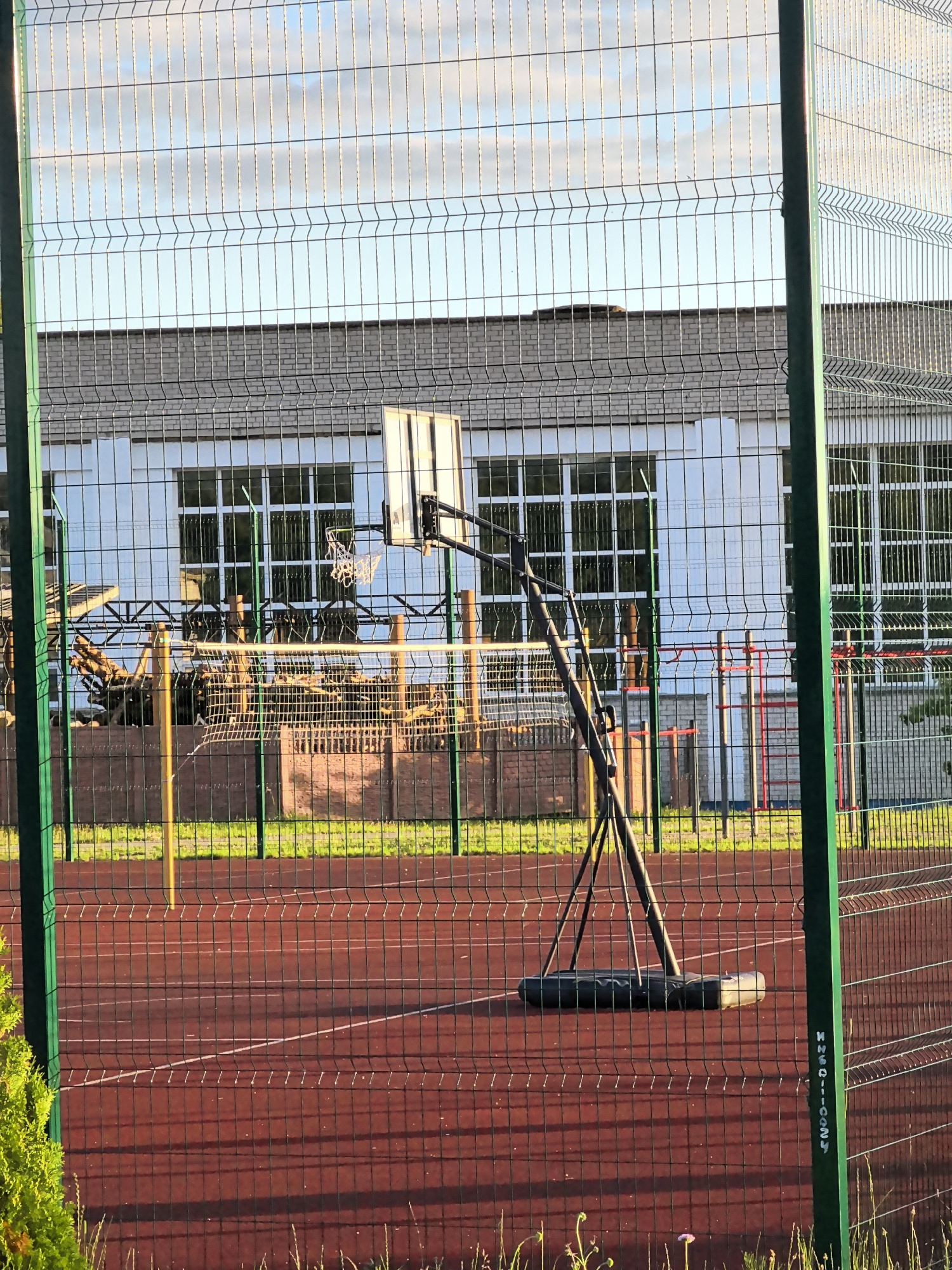 The image shows an outdoor basketball court with a portable basketball hoop in the foreground. The court is made of a red, textured material with white lines marking the boundaries. A green metal fence surrounds the court, with another fence in the background. Behind the fence, there is a building with white walls and large windows. The building appears to be a school or an educational facility. There are some playground structures visible behind the basketball hoop, including wooden logs and climbing equipment. The sky is partially cloudy, with a few scattered clouds visible. The lighting suggests it is either early morning or late afternoon, as the sun casts long shadows. There are no people visible in the image. The environment is clean and well-maintained, with green grass growing along the edges of the court.