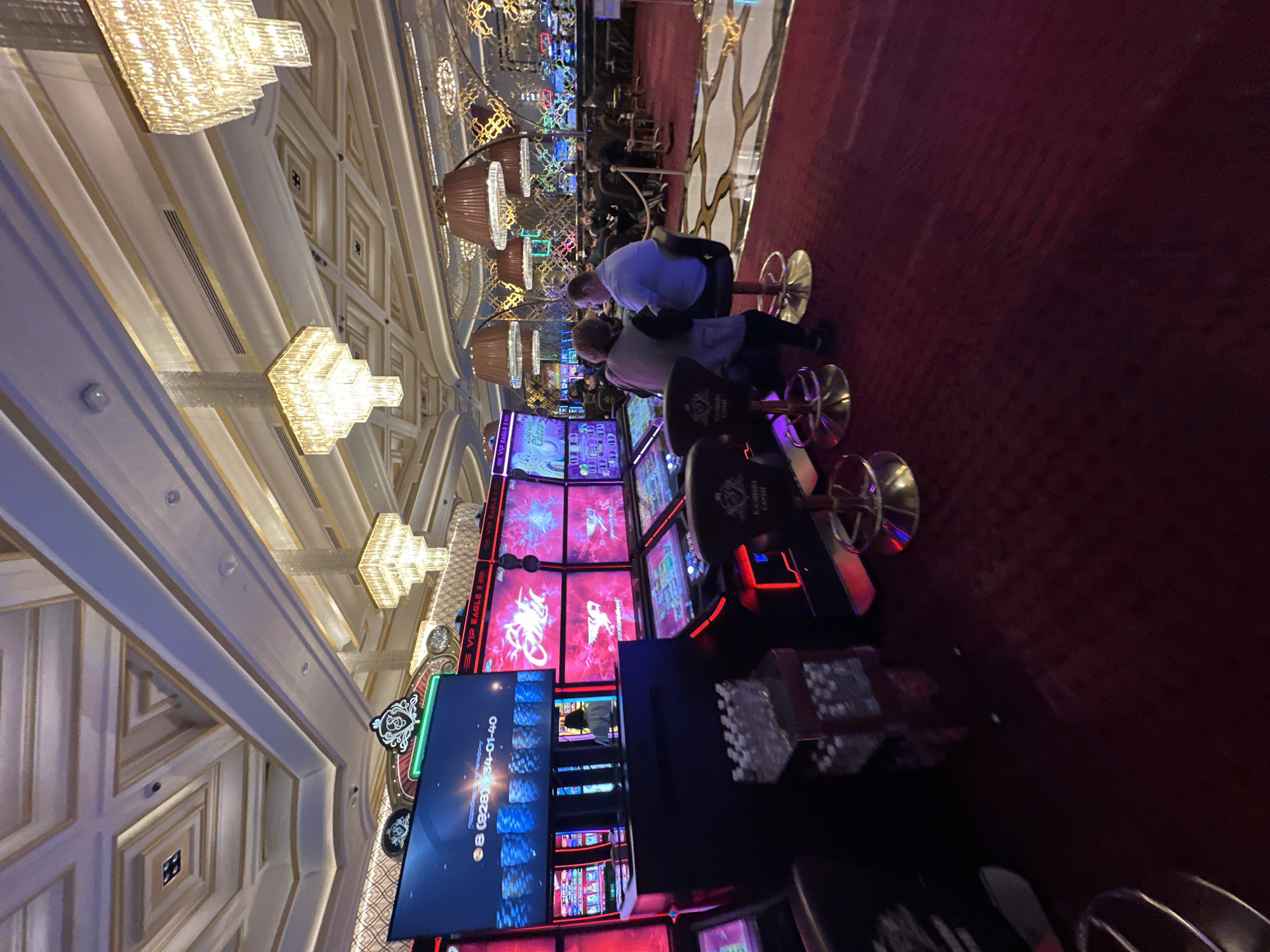 The image shows the interior of a casino, specifically the gaming floor. Several slot machines are visible, with their screens displaying various game graphics and information. Two people are seated at one of the slot machines, a man and a woman, appearing to be playing. In the background, other patrons and casino staff can be seen scattered throughout the venue, some seated at tables or standing. The casino is ornately decorated with plush red carpets, elaborate chandeliers, and gold accents on the ceiling and walls. The lighting is dim and atmospheric, typical of a casino environment. The scene appears to be set in a high-end establishment, suggesting a luxurious and immersive gaming experience.