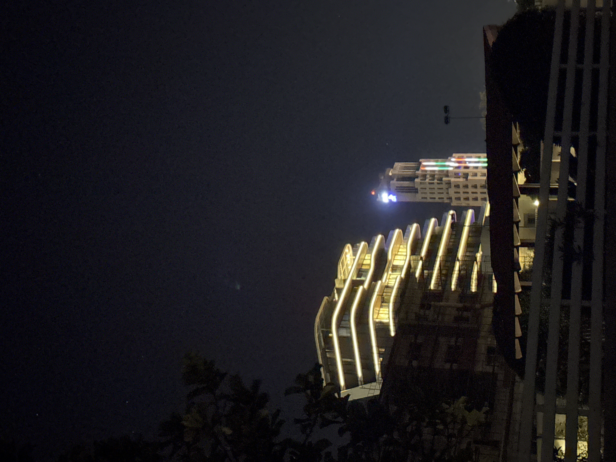 A nighttime exterior shot captures a multi-story building with cascading balconies, illuminated by horizontal bands of warm yellow light. Adjacent to it, a taller, more slender building stands, displaying vertical bands of colored lights in red, green, and white, topped with a blue illuminated structure. The scene is set against a dark night sky with a few faint stars visible. In the foreground, a white picket fence runs horizontally across the lower portion of the frame, partially obscuring a lower building and some shadowy foliage to the left. The overall impression is one of urban nighttime illumination. This scene is located in Parekklisia Community, Cyprus.