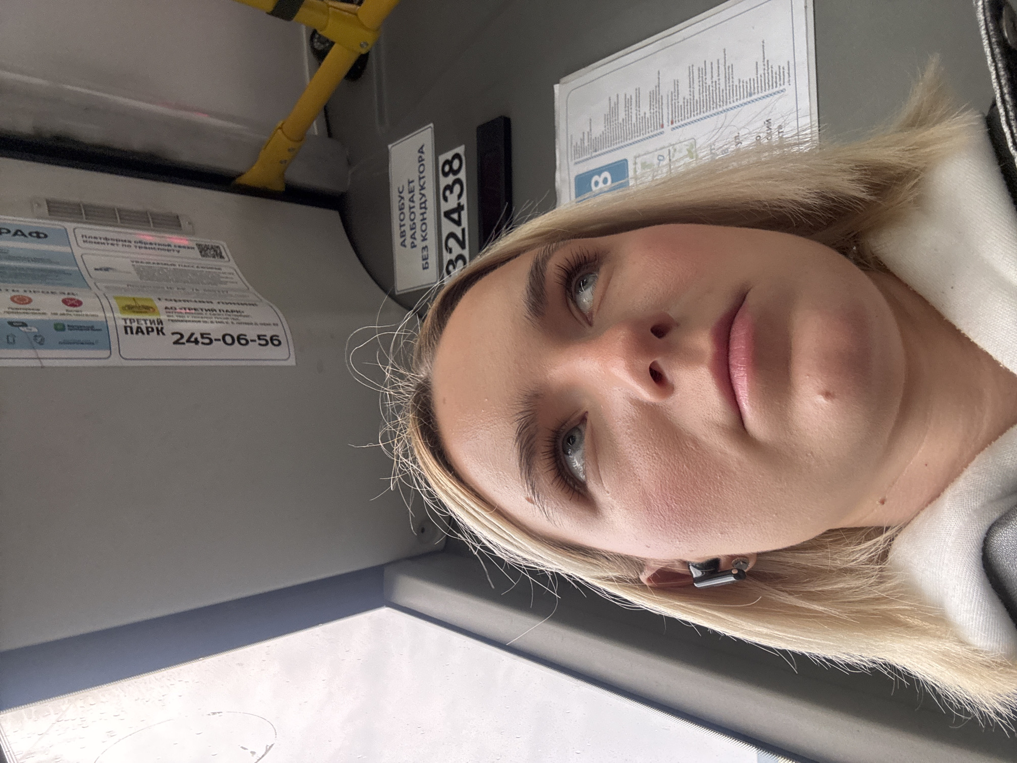 The image depicts a fair-skinned, blonde-haired woman inside a public transportation vehicle. An identification plate on the wall behind her has the number 32438. A sticker next to the plate says, "Autobus Rabotaet Bez Konduktora" indicating that the bus operates without a conductor. A sticker with Cyrillic text and the number 245-06-56 is also visible. The location is Vsevolozhsk, Russia.