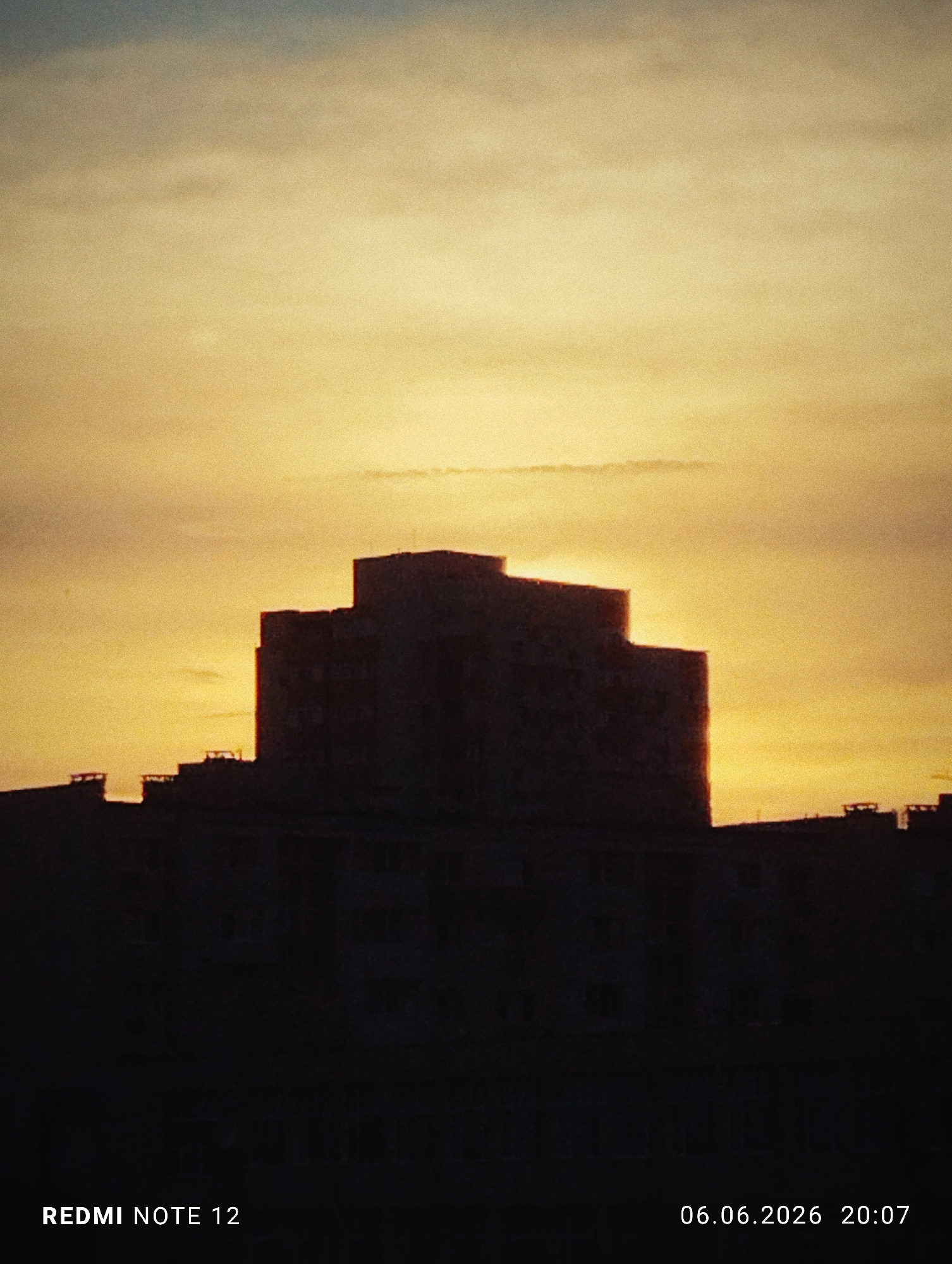 The image depicts a sunset scene over the city of Burgas, Bulgaria. The sky is dominated by a warm, golden-yellow light that transitions to a muted grey-blue tone at the very top. Wispy, indistinct clouds are scattered across the sky. In the midground, a prominent, multi-story building is silhouetted sharply against the bright sky, revealing a tiered or stepped roofline. To the left and right of this central structure, several other shorter buildings are also rendered as dark silhouettes. The foreground is completely underexposed, appearing as an opaque black mass with no discernible details. No people, specific actions, or interactions are visible in the scene. The image was captured with a REDMI NOTE 12 device on June 6, 2026, at 20:07, and exhibits a noticeable grain or noise texture.