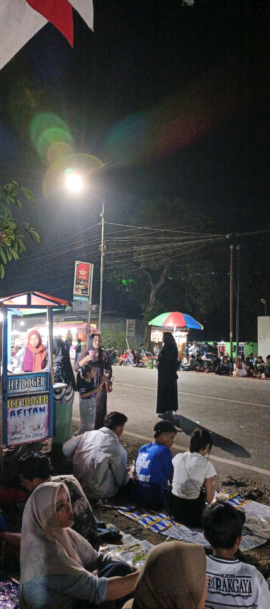At night in Kasiyan, Indonesia, people are gathered along the side of a road. In the foreground, several individuals are seated on the ground, some on mats. A food cart selling "ICE DOGER" is visible on the left, with a woman in a hijab standing behind it. Further down the road, a person in a black abaya stands under a colorful umbrella. Other people are seated on the ground across the road, some near small tables and stalls. A street lamp illuminates the scene, casting long shadows. The atmosphere appears to be that of a local gathering or event.