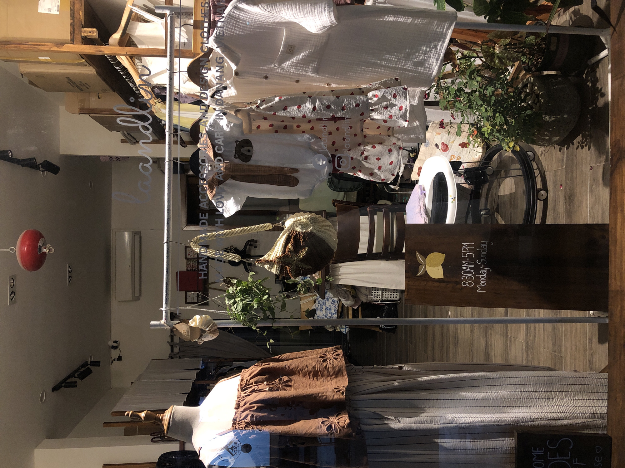 This image showcases the interior of a retail shop, likely a boutique, displaying a variety of clothing and accessories. The scene is artfully arranged, with garments hanging on a rack and a mannequin draped in clothing. Among the displayed items are what appear to be children's outfits, one featuring a strawberry pattern and another with a bear appliqué. Other items include woven bags and a white linen shirt.
Prominently visible on a wooden sign is text indicating the shop's hours of operation: "8:30AM-5PM Monday-Sunday". Above this, another sign reads "HANDMADE ACCESSORIES AND DESIGN CLOTHES" and suggests a location: "IN DANANG". There's also text that appears to say "HOI AN" and possibly "HOI AN DES", hinting at a connection to the nearby historic town.
The lighting suggests it is daytime, with natural light illuminating the display. The overall impression is one of a curated collection of unique, possibly handmade, items presented in a visually appealing manner. The presence of plants adds a touch of natural ambiance. The shop appears to be located in a bustling area, given the mention of Đà Nẵng.