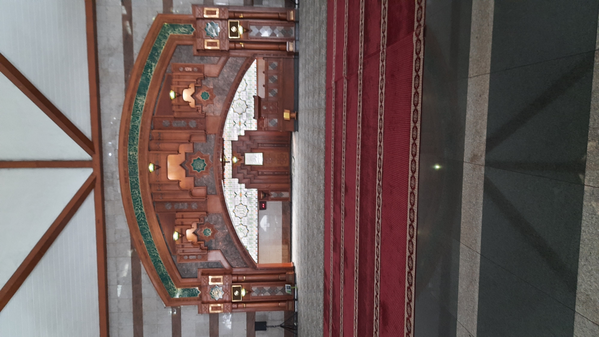 The image shows the interior of a mosque, likely in Bandung City, Indonesia, featuring a large wooden mihrab with intricate carvings and Islamic calligraphy. The mihrab is adorned with green accents and illuminated by several lights. In front of the mihrab is a red carpet with a patterned border, laid out in prayer rows. The floor is a combination of dark and light colored tiles. The ceiling is supported by wooden beams, and the walls are paneled with beige and brown tiles. There are no people visible in the image. The overall scene depicts a well-maintained and ornate place of worship.