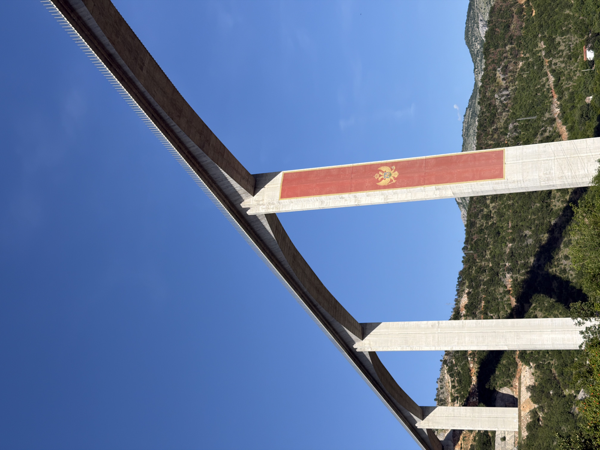 The image displays a large concrete viaduct, located in Gornje Mrke, Montenegro, viewed from a low-angle perspective looking upwards. The structure consists of multiple tall, slender concrete piers supporting a broad, flat bridge deck. One prominent pier, positioned towards the center-right of the frame, features a large, vertically oriented rectangular banner painted in red with a golden border. Centered on this red banner is the golden Coat of Arms of Montenegro, depicting a double-headed eagle clutching an orb and scepter, with a lion on its chest shield. The top edge of the bridge deck is lined with a series of white, spike-like elements. The viaduct spans across a valley characterized by steep, undulating hillsides densely covered in various green vegetation and trees. The background shows distant mountains under a clear, vibrant blue sky. Distinct shadows are cast by the bridge piers and the terrain, indicating bright sunlight. A small white building with a red roof is discernible at the base of a distant hill on the lower right side. There are no people, vehicles, or animals visible in the scene.
