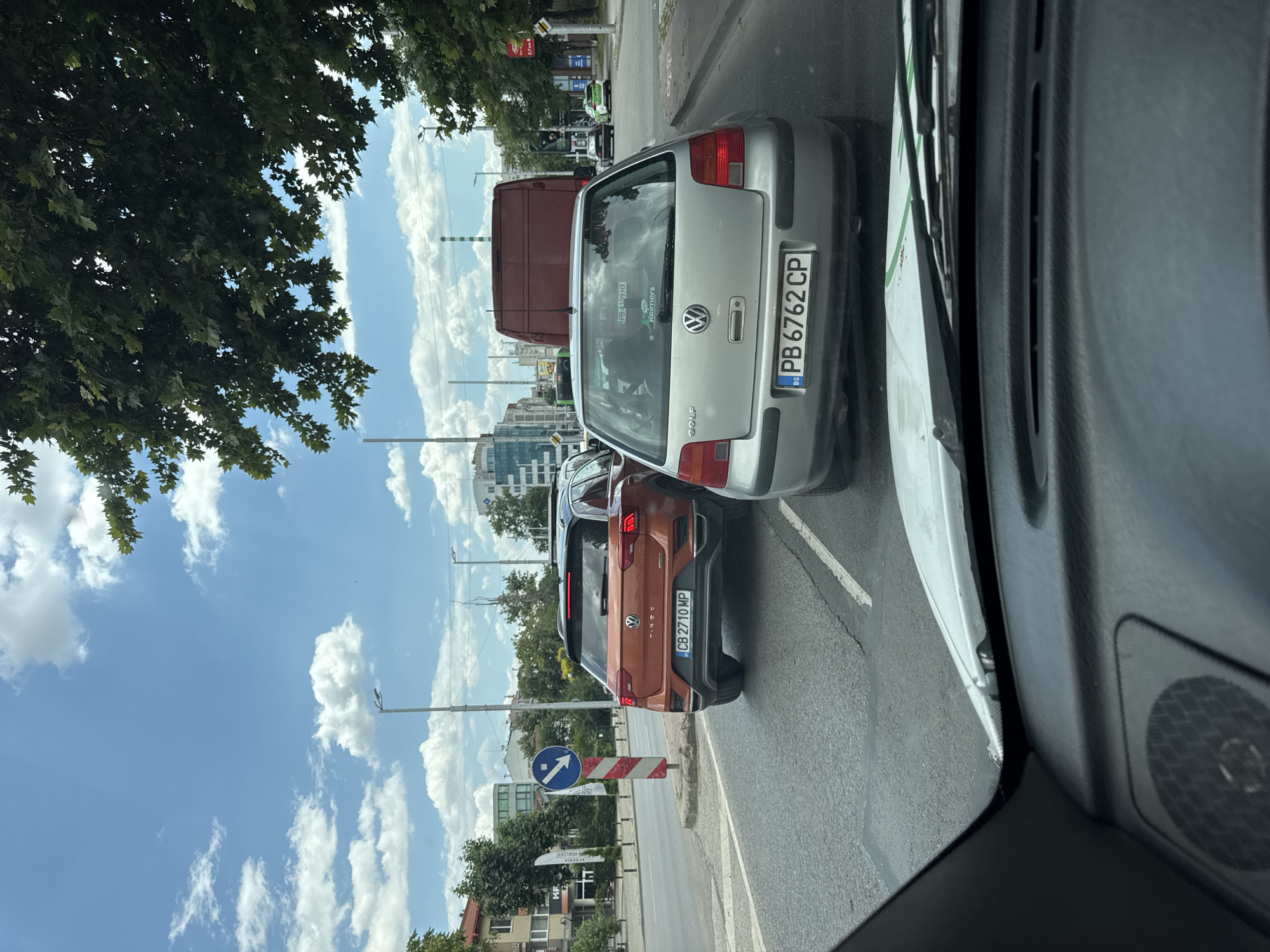 The image depicts a traffic scene in Plovdiv, Bulgaria. The foreground shows the interior of a vehicle, with part of the dashboard and windshield visible. Outside the vehicle, several cars are stopped in traffic, including a silver Volkswagen Golf with the license plate PB 6762 CP. Behind the Golf is a large, reddish-brown van, and further ahead, an orange Volkswagen T-Roc with the license plate CB 2710 MP is visible in the adjacent lane. The background features buildings, trees, streetlights, and a blue sky with white clouds. A traffic sign with a blue circular arrow indicating a left turn is positioned on the side of the road. The overall environment suggests an urban setting during daylight hours.
