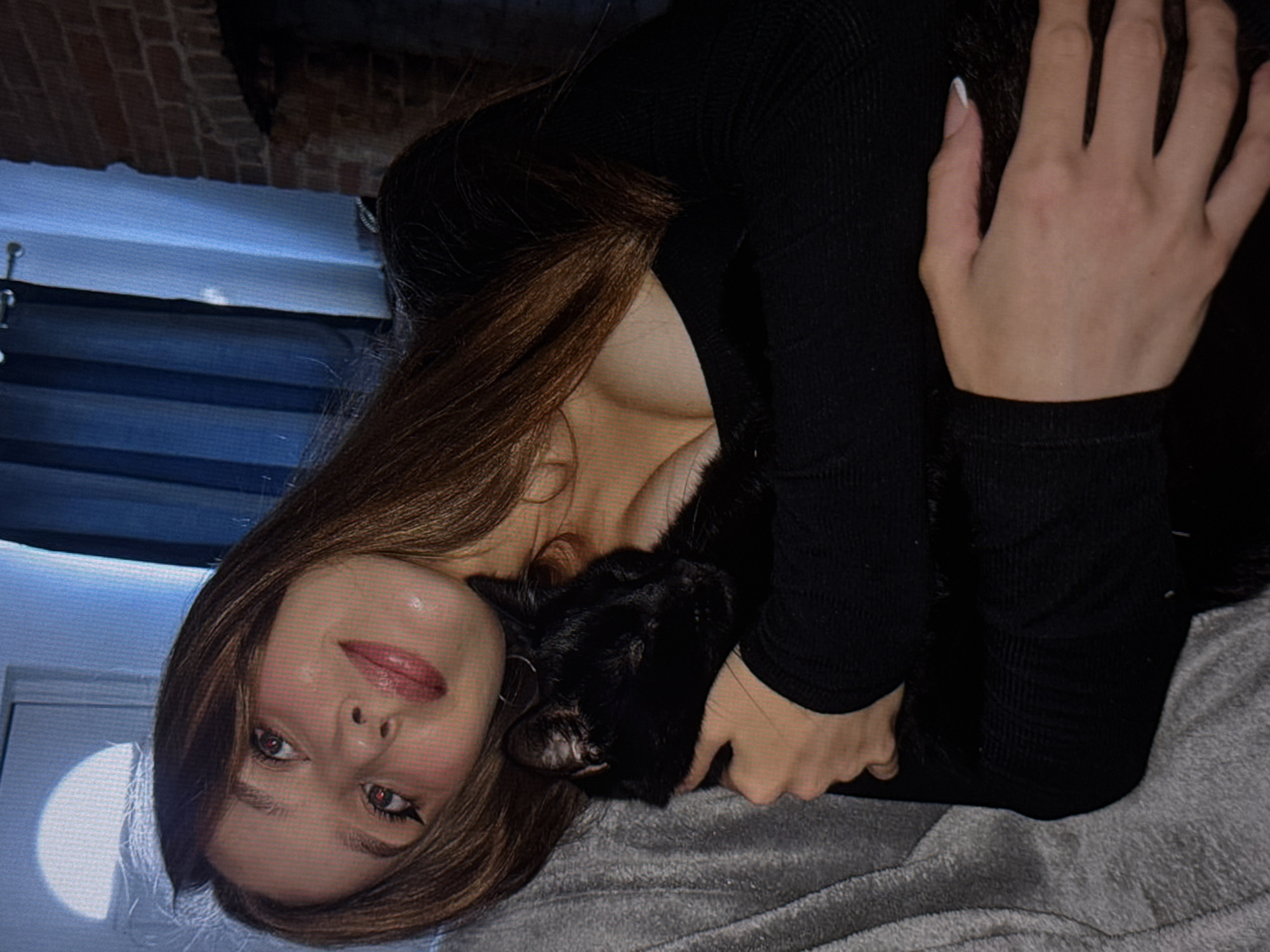 A young woman with long, brown hair is seen from the chest up, holding a black cat. She is wearing a black, long-sleeved shirt with a low neckline. The woman is lying down, with her head tilted back and to the side, and her eyes are partially closed. The cat is nestled in her arms, and its eyes are also closed. The background of the image is blurred, but it appears to be an indoor setting with a window and curtains. The lighting is dim, creating a cozy and intimate atmosphere.