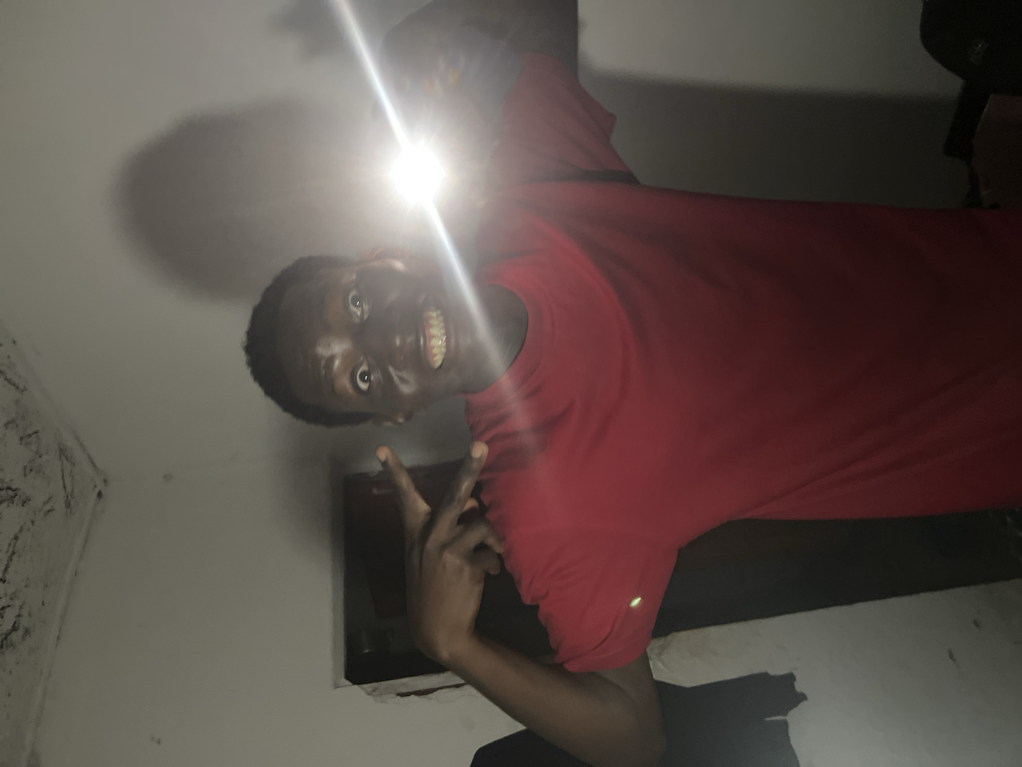 In a dimly lit, possibly residential room in Agwala, Nigeria, a young Black male is captured in a close-up, waist-up shot. He is wearing a bright red t-shirt and has a wide, somewhat startled expression on his face, with his eyes open and teeth bared in a grimace. A bright light, likely from a flash or phone, is directed towards the camera, causing lens flare and obscuring part of his face and the background. His left arm is raised, and his fingers are positioned in a gesture that appears to be a "rock on" sign, with his thumb extended and index and pinky fingers raised. The background shows a rough, textured wall and ceiling, suggesting an unfinished or aged interior. A dark opening, possibly a doorway or niche, is visible to the left of the subject. The overall impression is one of a spontaneous, possibly playful, but also somewhat unsettling photograph taken in a basic indoor environment.