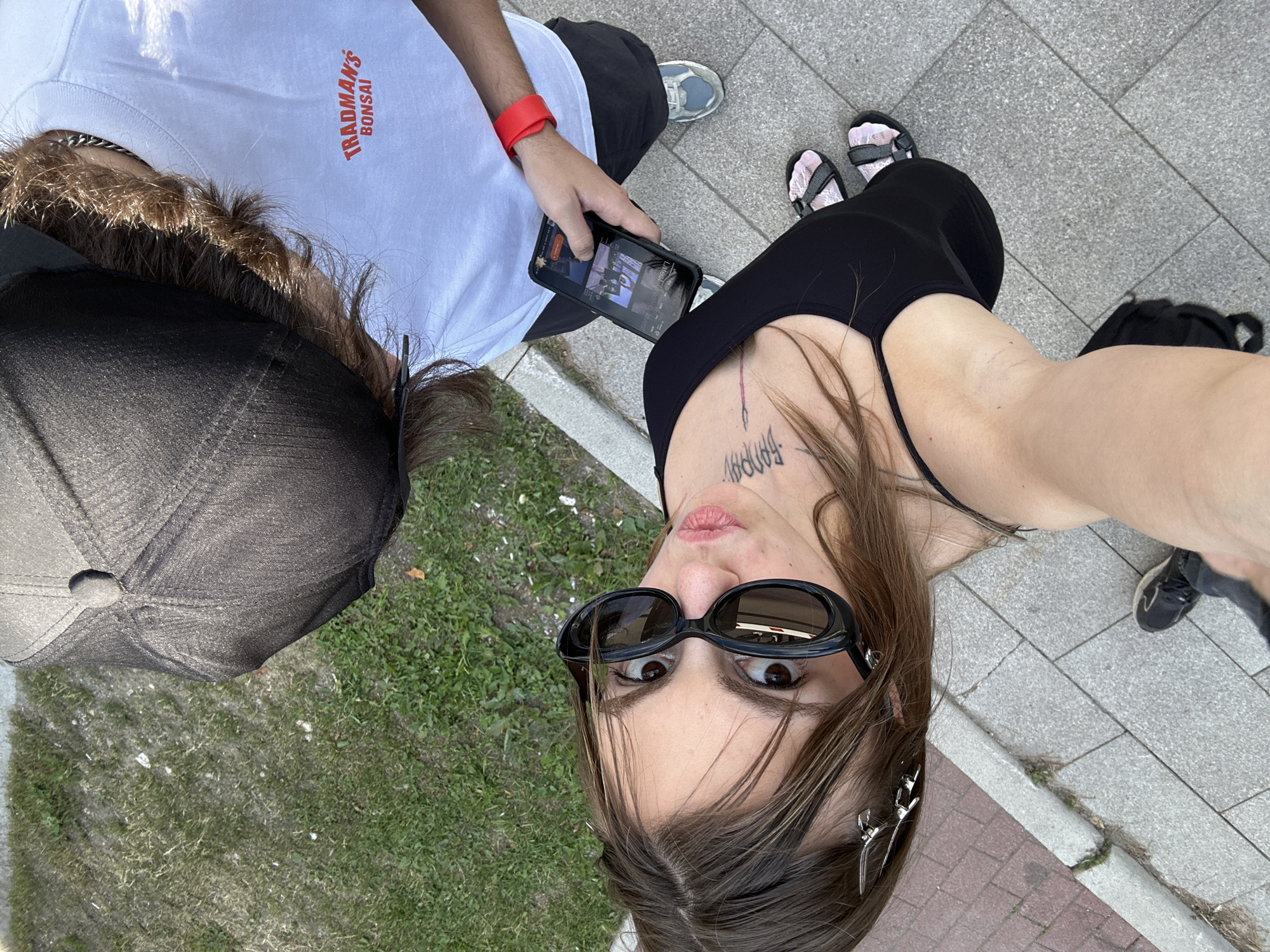 Two individuals are captured in a high-angle, selfie-style photograph taken outdoors on a paved sidewalk adjacent to a grassy area. The person in the foreground, on the left, is a woman with long, brown hair, wearing large, black sunglasses, a black tank top, and pink and gray sandals. She has tattoos on her neck and chest. The second individual, partially visible on the right, appears to be wearing a white t-shirt with red text that reads "TRADMAN'S BONSAI," a black baseball cap, and athletic shoes. This person is holding a smartphone, displaying an image, and is wearing a red wristband. The ground is a mix of grey paving stones and a strip of red brick border. The environment suggests a public outdoor space, possibly a park or a street in Moscow, Russia, given the context of the prompt. The lighting indicates it is daytime.