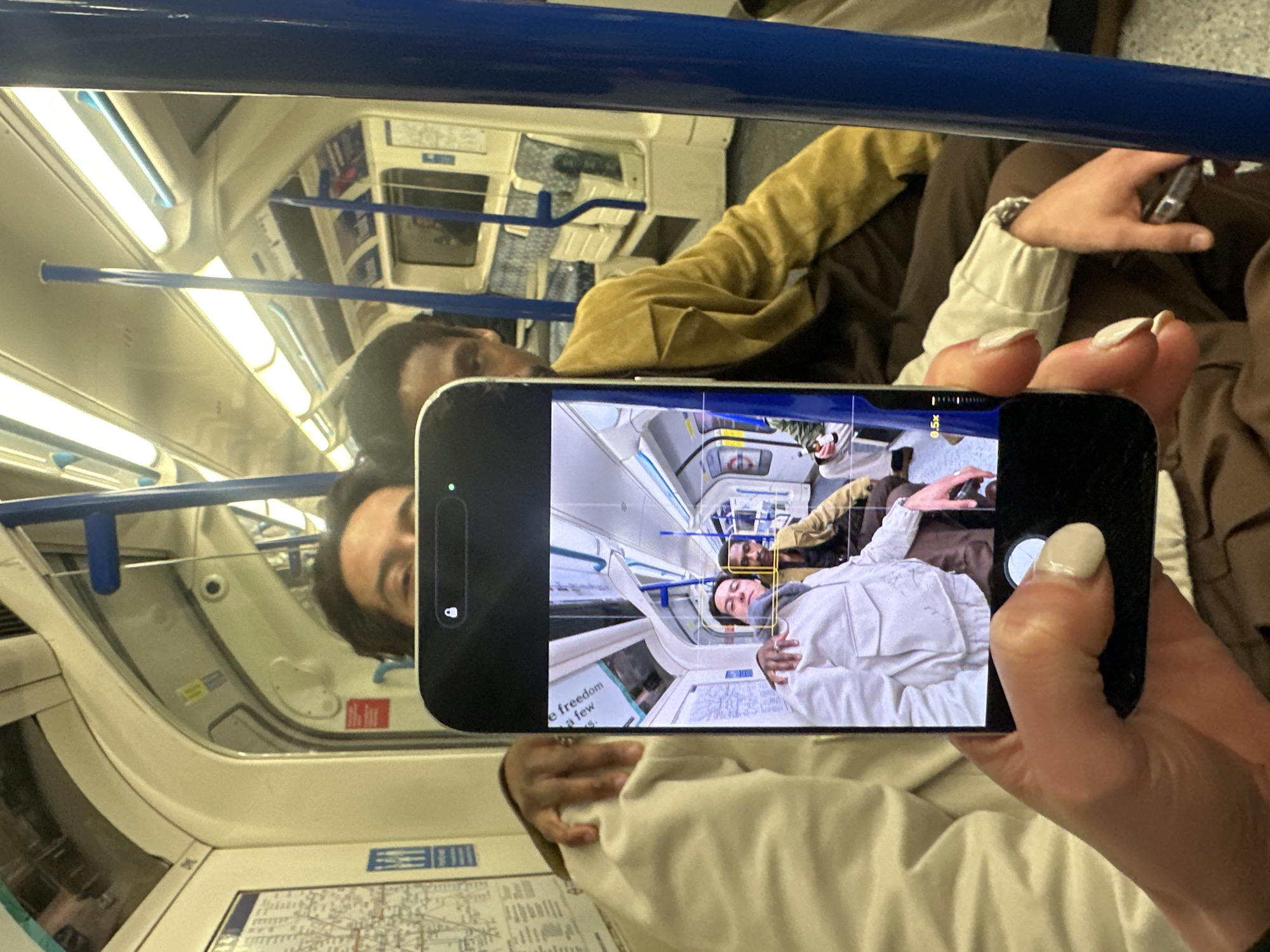 A mirror selfie is being taken inside a London Underground train carriage. The camera is held by a person whose right hand, with light-colored manicured fingernails, is visible depressing the shutter button on a black smartphone. The phone screen displays a reflection of two seated males. The male on the left in the reflection wears a cream jacket with two chest pockets and has his right arm around the other male's shoulder. The male on the right in the reflection wears a mustard yellow jacket and has his left arm around the first male's shoulder. Both males are visible in full in the phone's reflection, and their heads are partially visible above the phone itself in the larger image. The train interior features white curved panels, blue vertical support poles, fluorescent ceiling lights, and patterned blue and white seating. A London Underground map is affixed to the wall on the left.