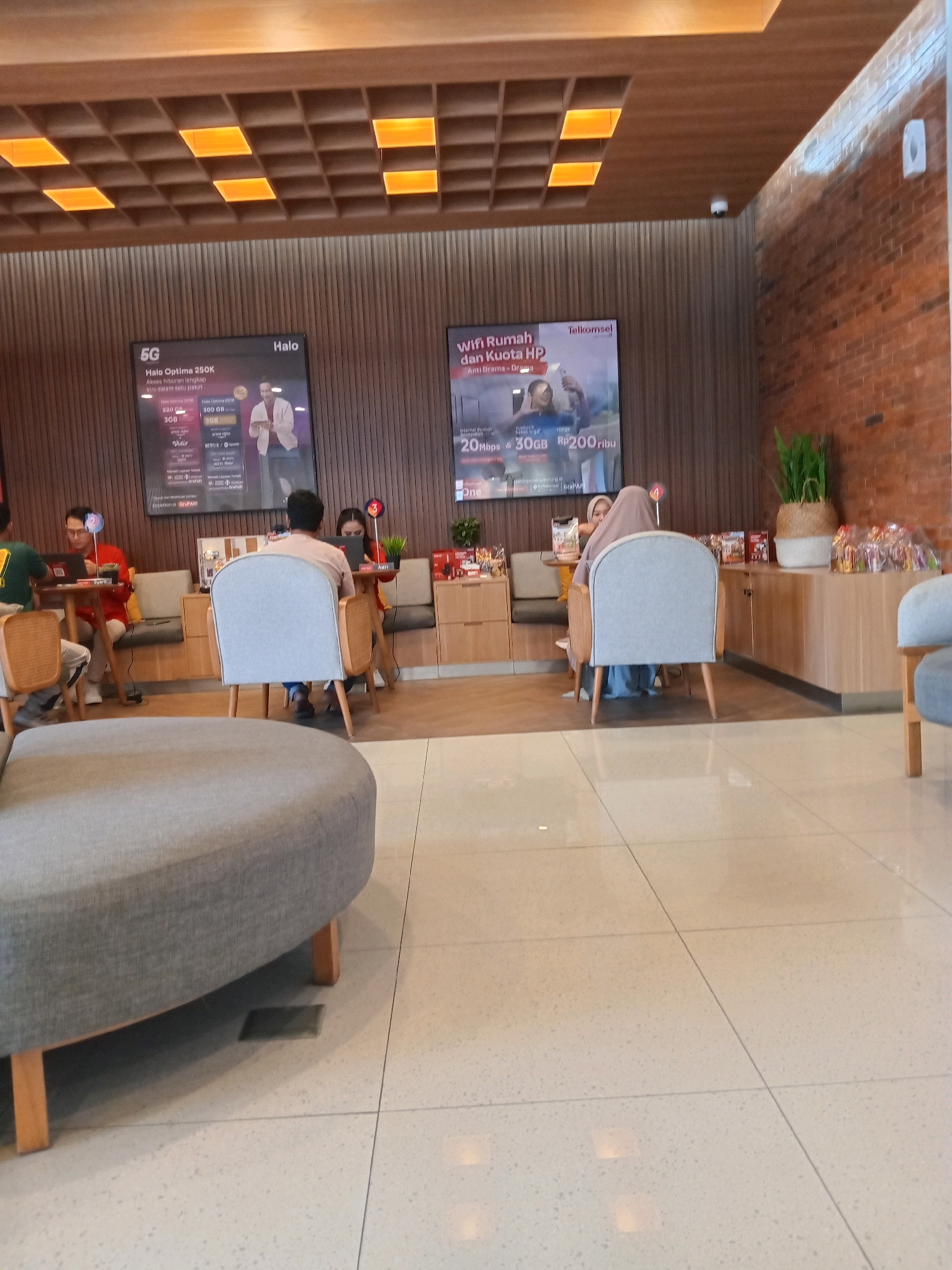 The image shows the interior of a Telkomsel store in Karangturi, Indonesia. The scene is well-lit, with wooden paneled walls and a grid-like ceiling with illuminated squares. In the foreground, a large, grey, curved sofa occupies the lower left of the frame. The middle ground features several seating areas with light-colored chairs and wooden tables.
In one seating area, a person wearing a green shirt is seated at a table with a laptop. To their right, another person in a red shirt is also seated at a table with a laptop. Further back, two individuals are seated at a round table. One is wearing a light-colored headscarf and a blue outfit, while the other, facing away from the camera, is wearing a light-colored long-sleeved shirt.
On the wall behind the seating areas, two large digital displays are visible. The left display advertises "Halo Optima 250K" and "5G", and the right display promotes "WiFi Rumah dan Kuota HP" with Telkomsel branding.
The background shows a counter with various items, including what appear to be product displays and a potted plant. The floor is tiled with large, light-colored tiles. The overall atmosphere suggests a modern and comfortable retail environment.